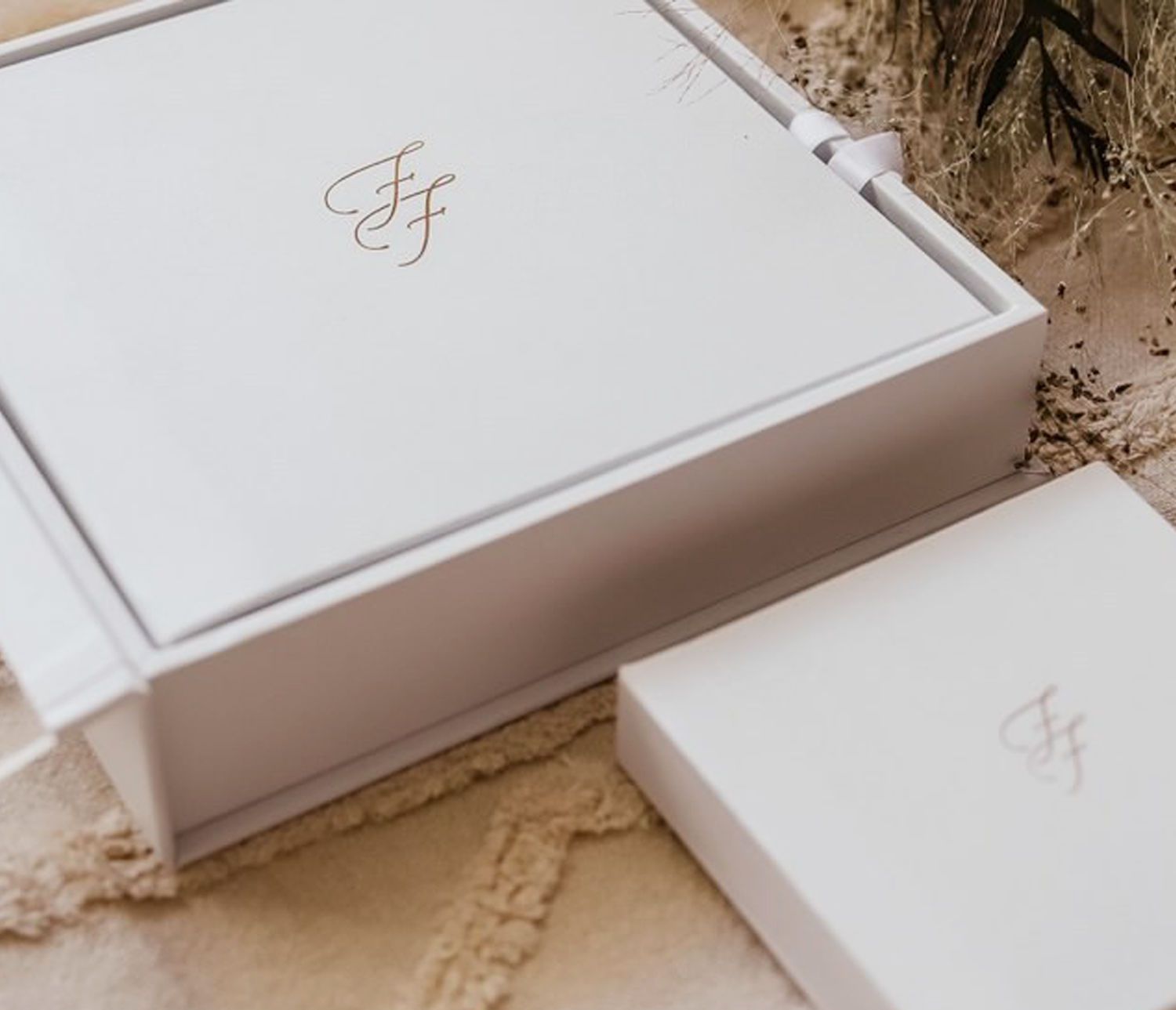 Two elegant white gift boxes with gold monogram initials displayed on a textured cream surface with dried botanicals.
