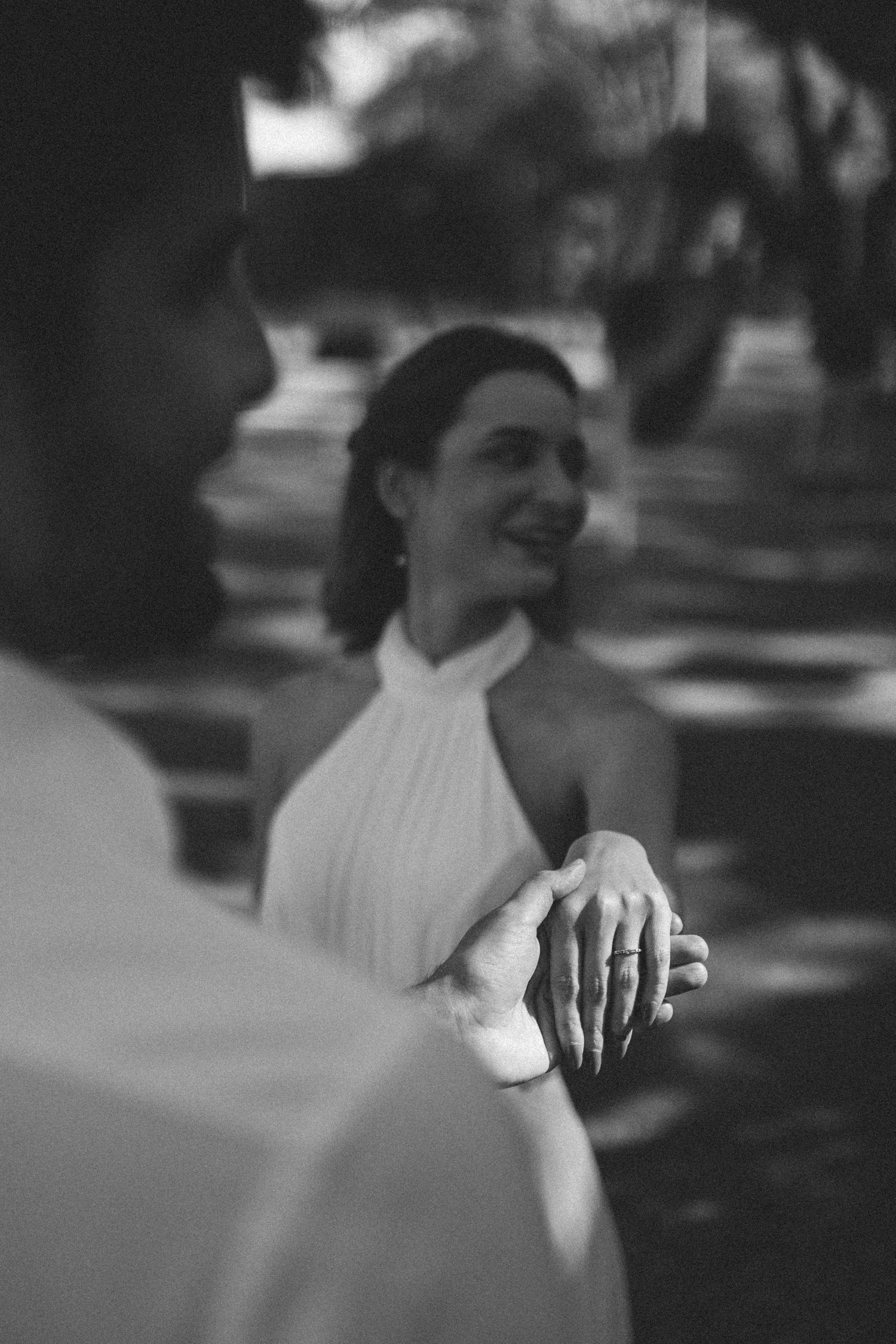 Black and white photo of smiling bride in halter dress holding hands with groom outdoors.