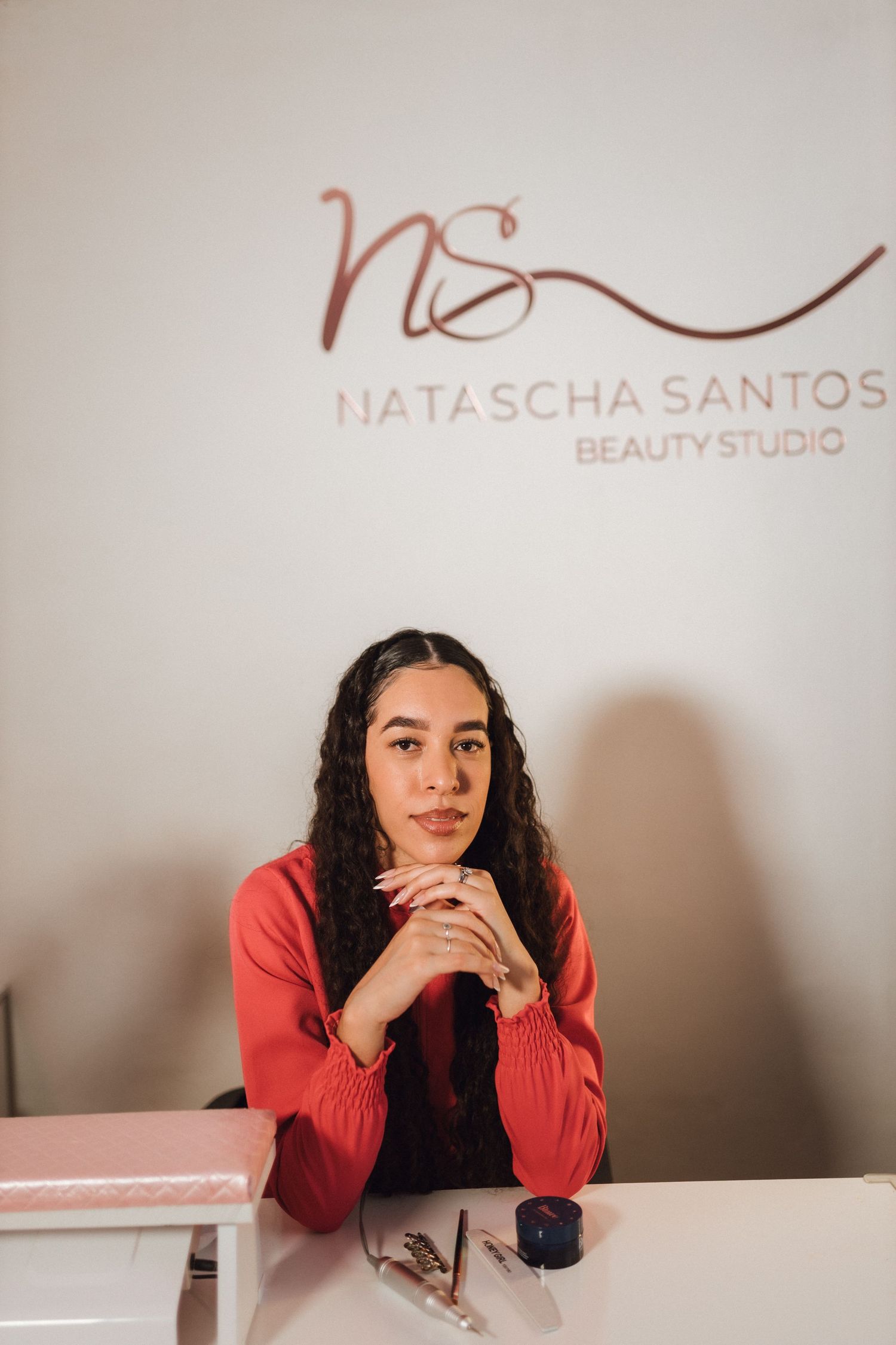 Woman in red blouse sitting at desk beneath Natascha Santos Beauty Studio sign.
