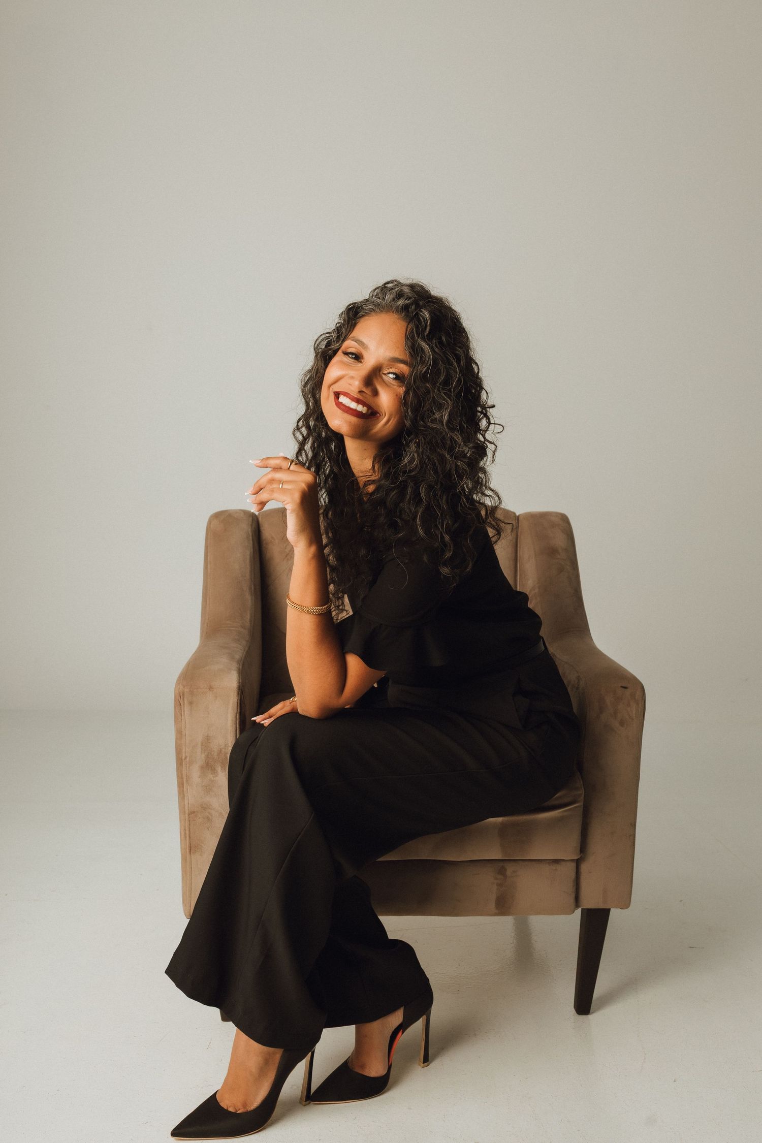 Smiling woman with curly hair in black outfit sitting in armchair.