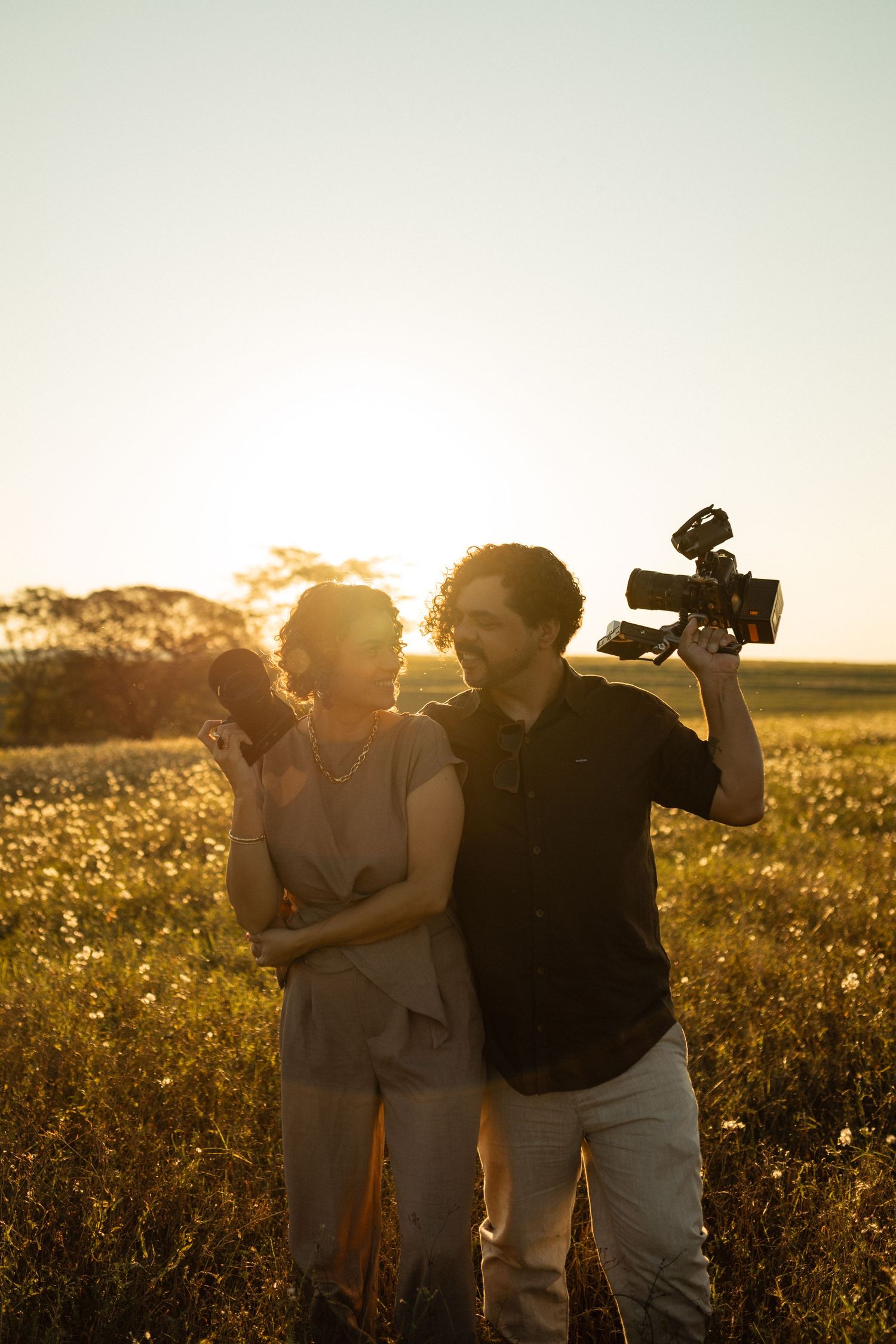 Two filmmakers stand in a golden sunlit field at sunset, man holding professional video camera aloft, woman smiling beside him.
