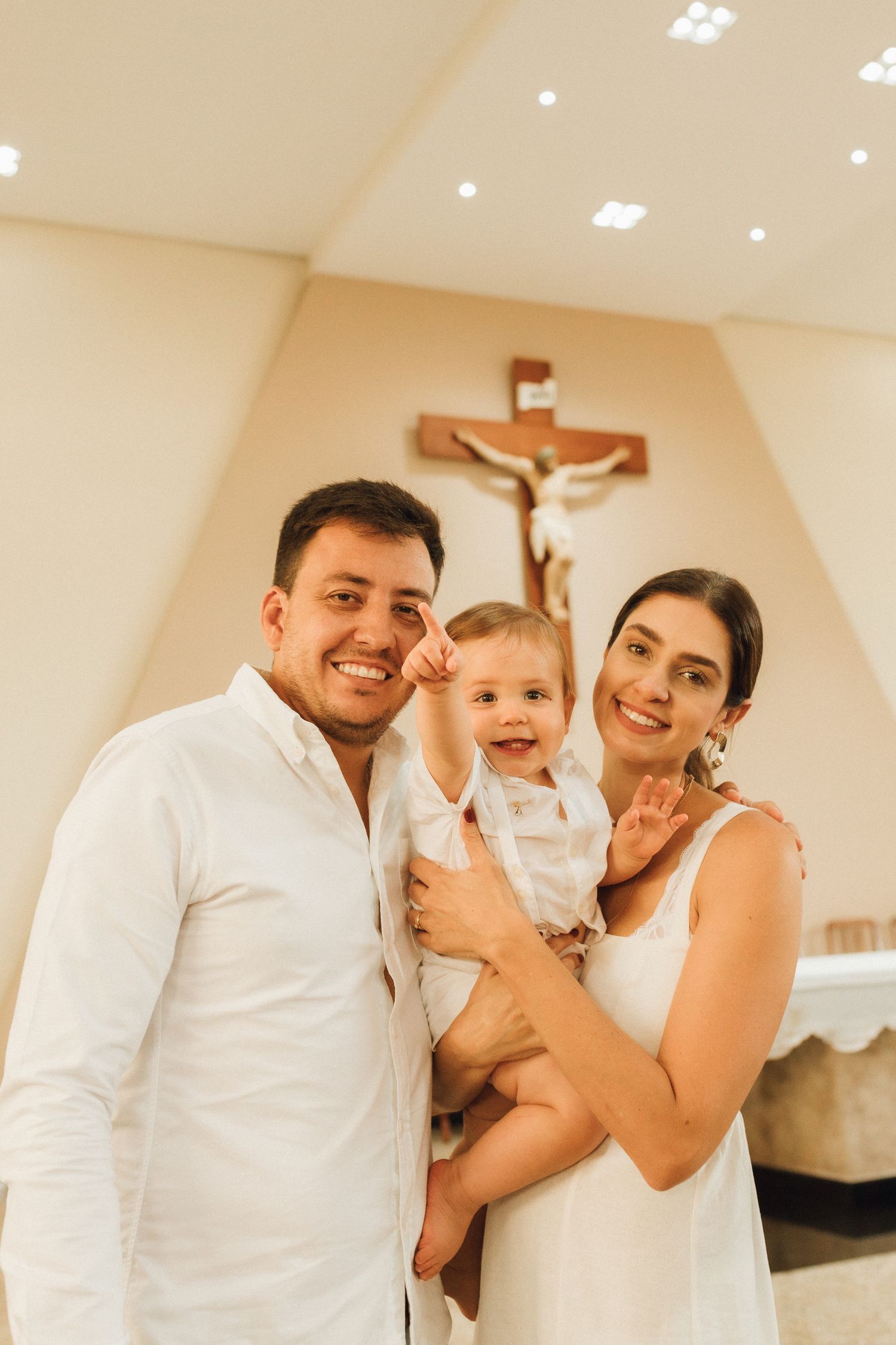 Happy family posing in church with crucifix, parents in white holding smiling baby at baptism ceremony.
