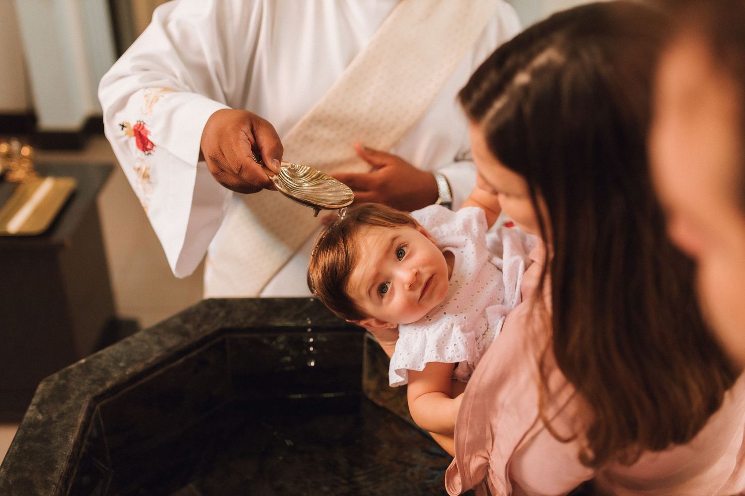 Priest baptizing a baby girl with holy water at a church font during a christening ceremony.