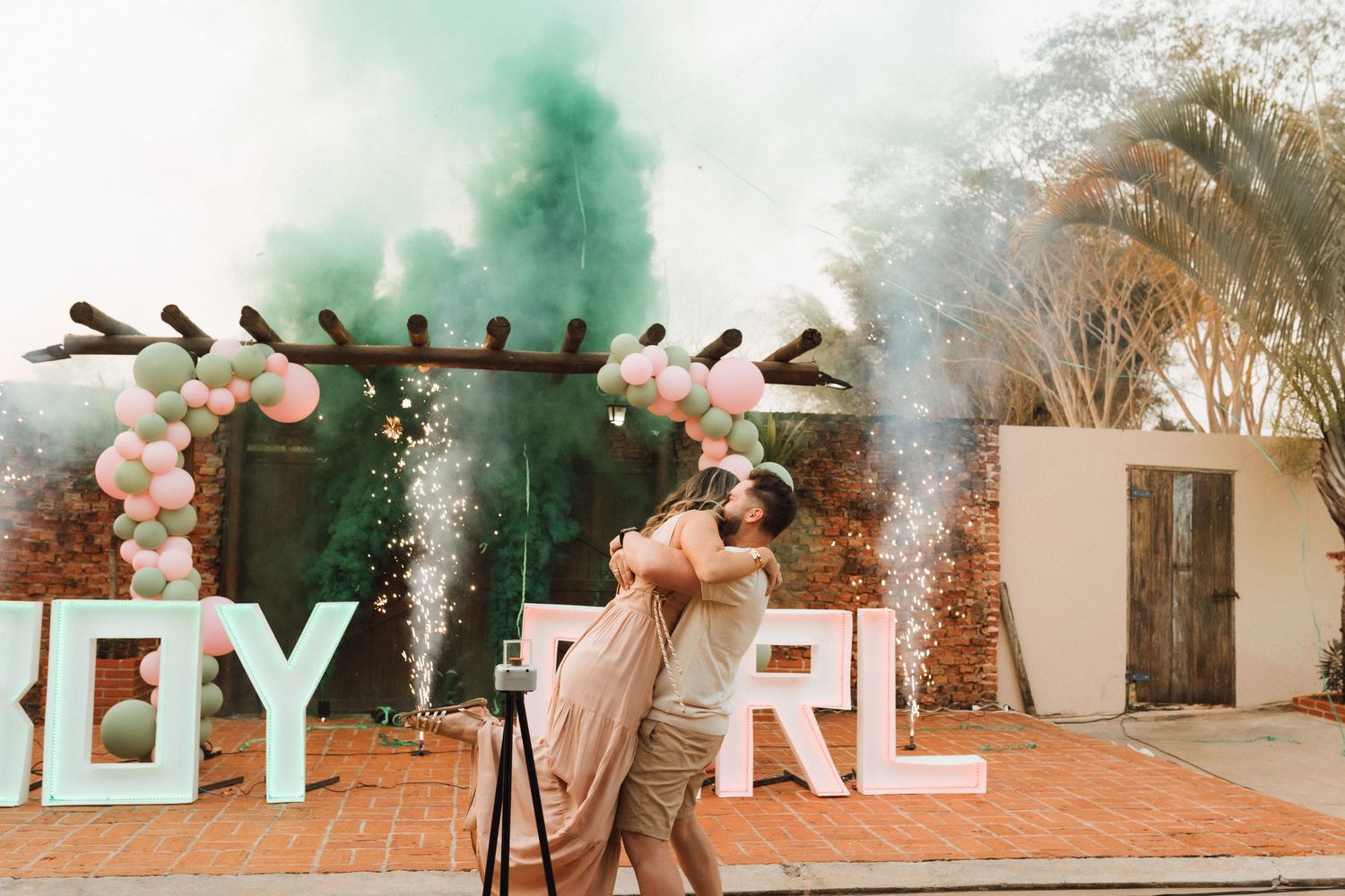 Couple embracing joyfully at gender reveal party with green smoke, balloon arch, and BOY GIRL letter signs.