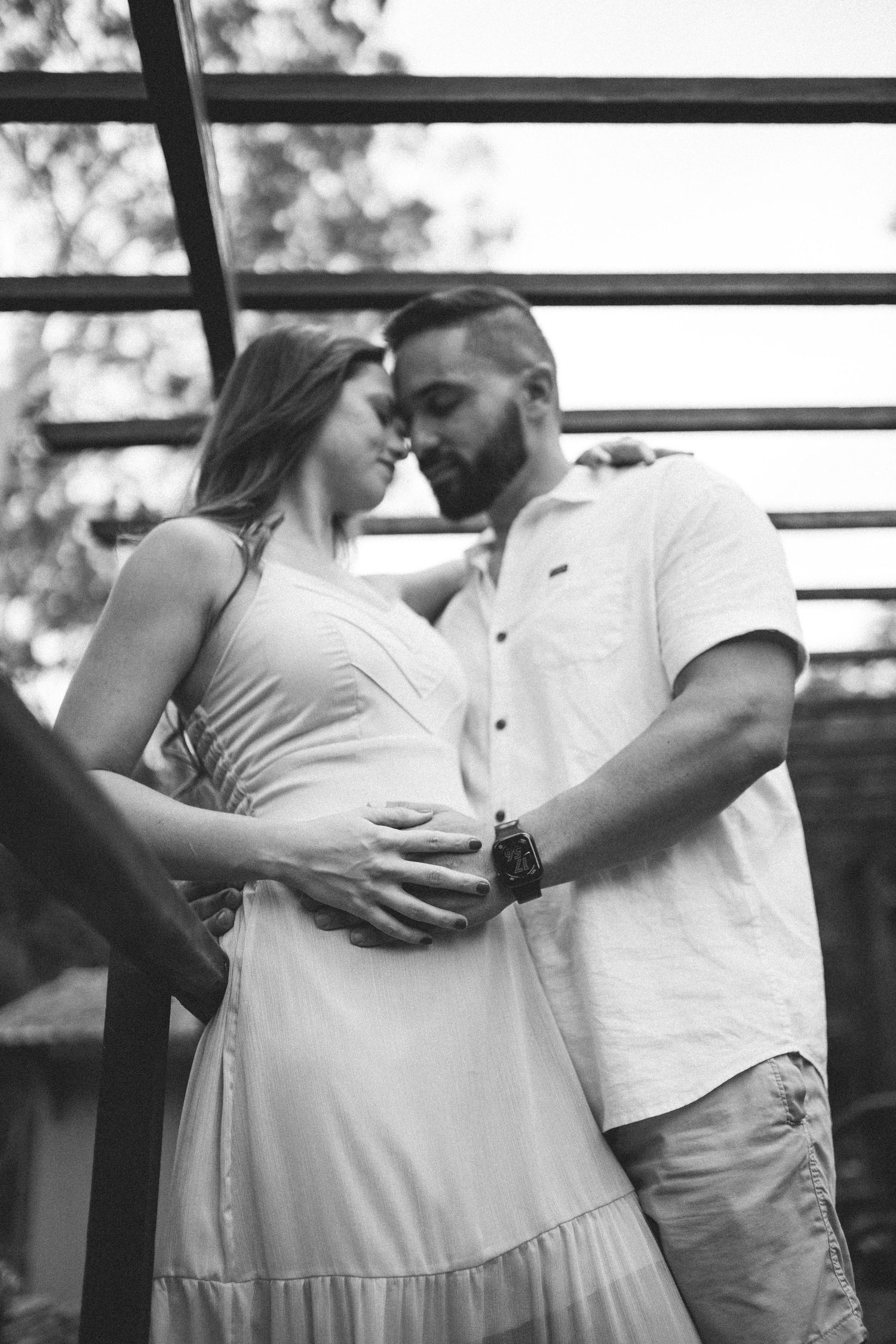Black and white photo of a couple tenderly touching foreheads, man cradling pregnant womans belly outdoors.