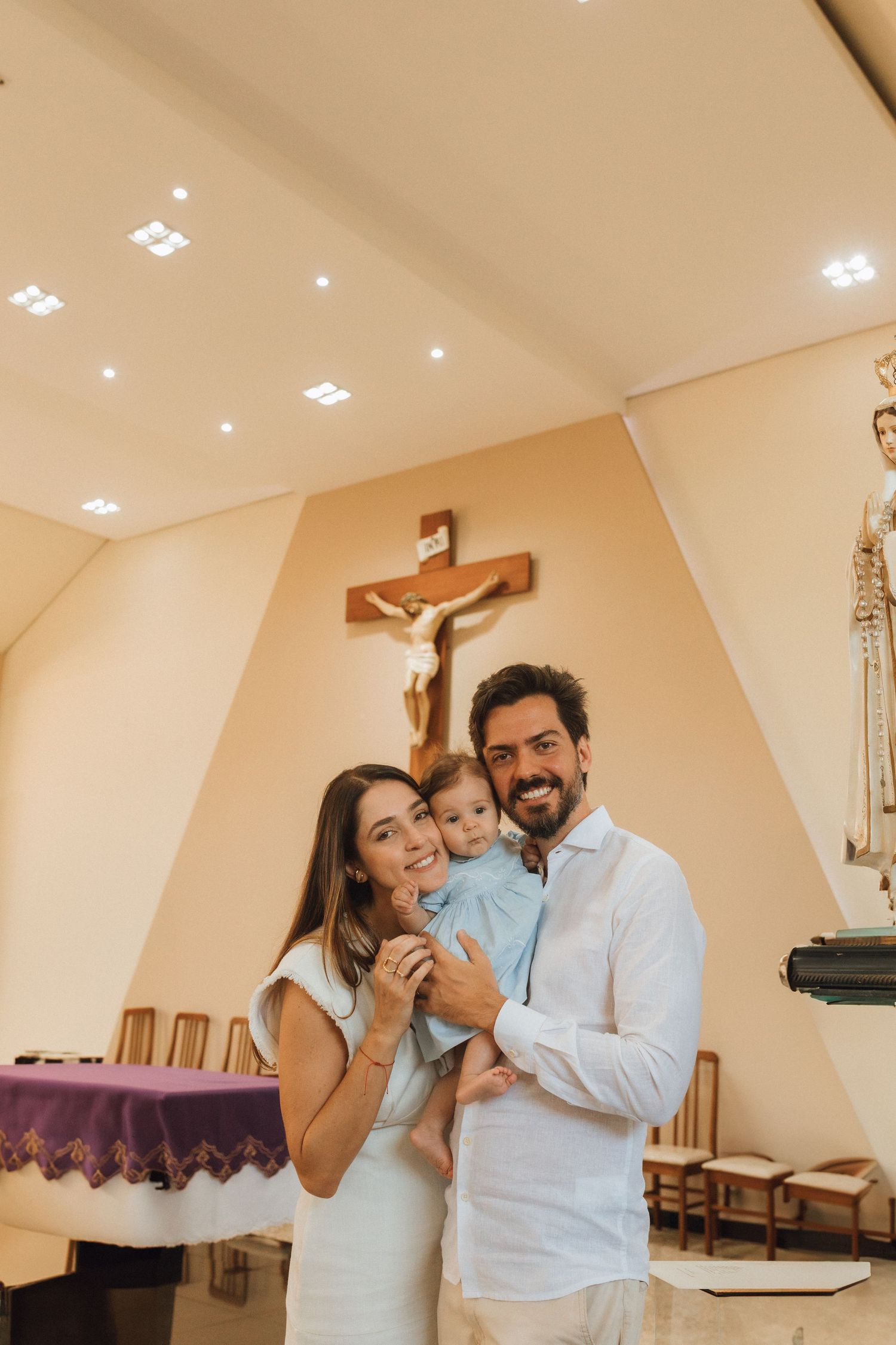 Happy family posing in a Catholic church with a wooden crucifix on the wall, parents holding their baby.