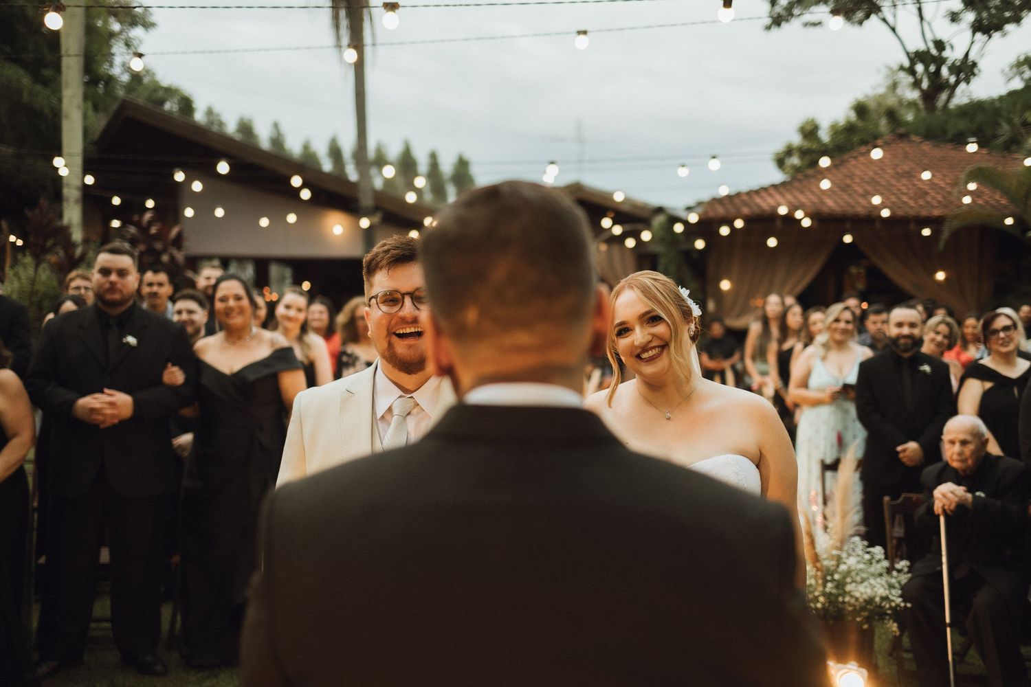 Groom laughing joyfully as bride smiles during outdoor evening wedding ceremony with string lights.