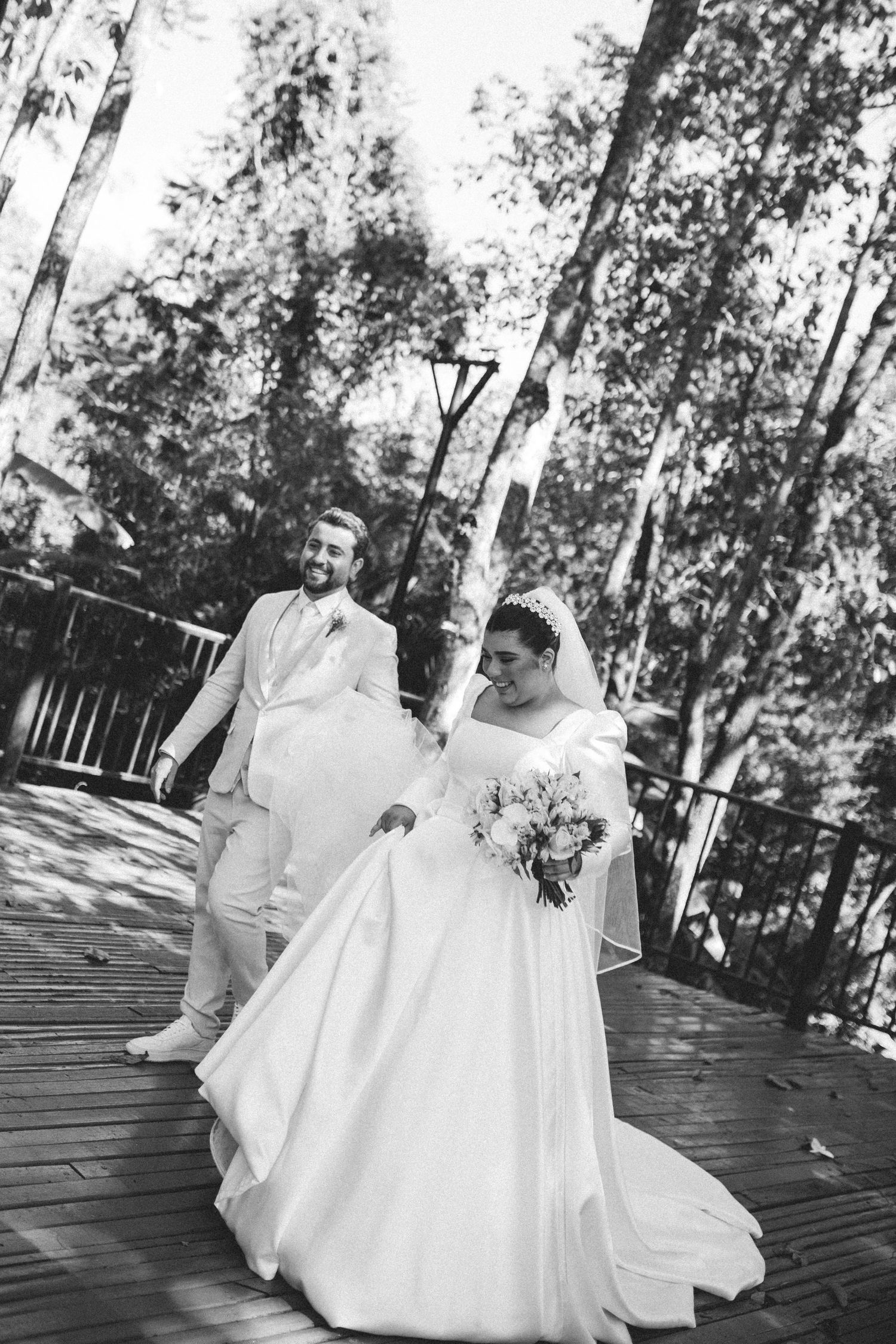 Black and white photo of a bride and groom walking hand in hand on a wooden deck surrounded by trees.