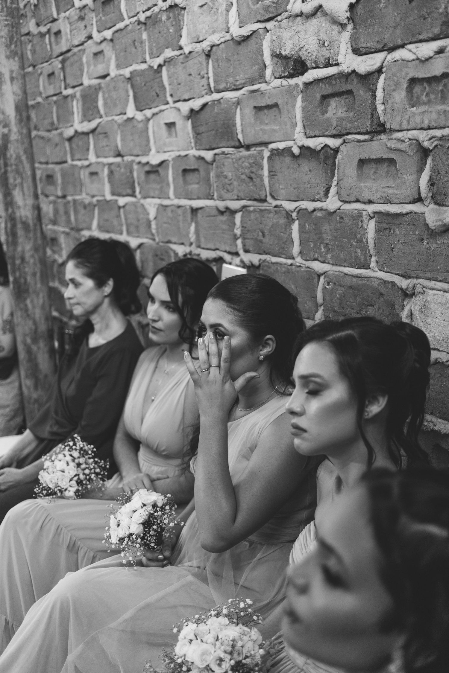 Emotional bridesmaids in pastel gowns sit against brick wall holding white bouquets during wedding ceremony.