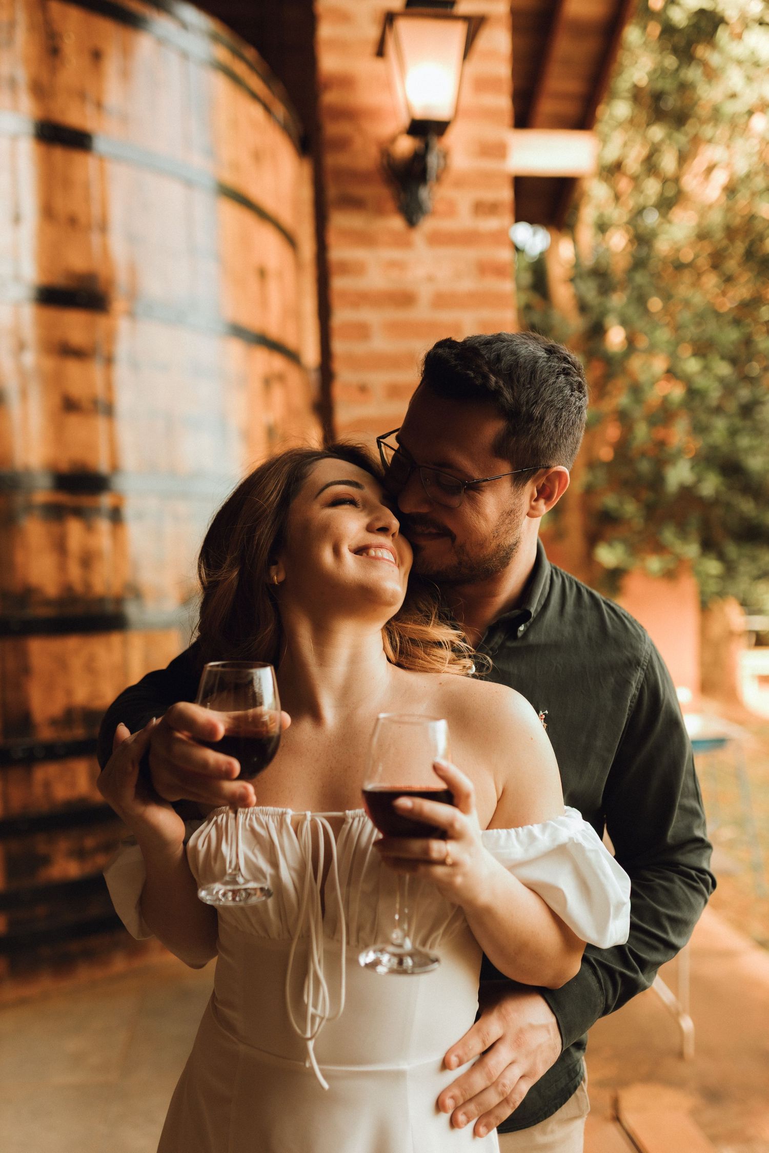 Romantic couple sharing wine glasses at a rustic winery with wooden barrels, man embracing woman from behind.