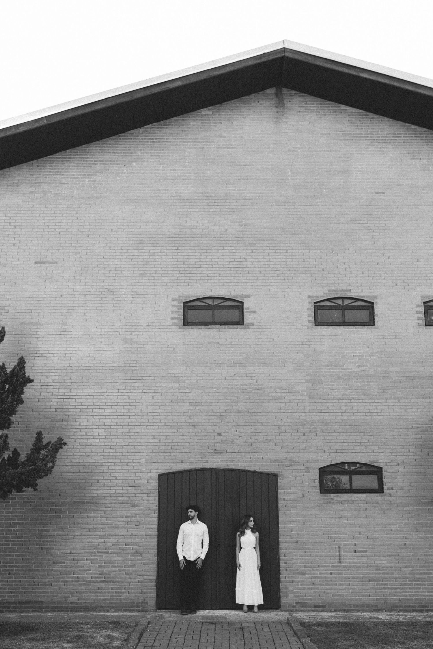 Black and white photo of couple standing at large barn doors of brick building.