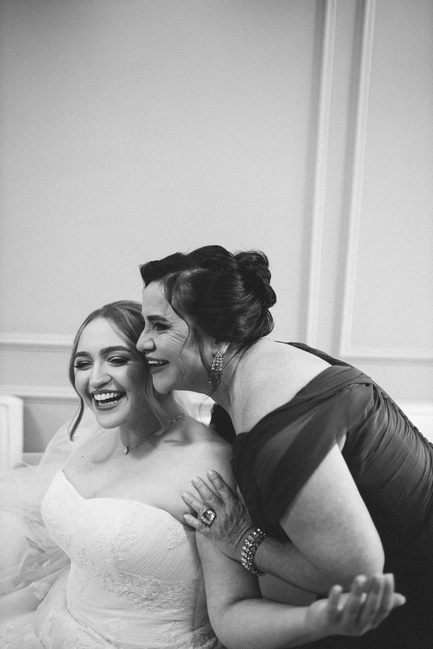 Black and white photo of laughing bride being embraced and kissed on cheek by female guest.
