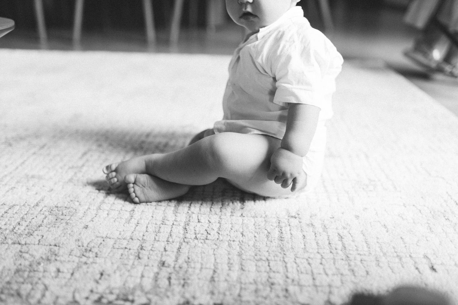 Black and white photo of a baby sitting on a carpet wearing a diaper and white shirt.