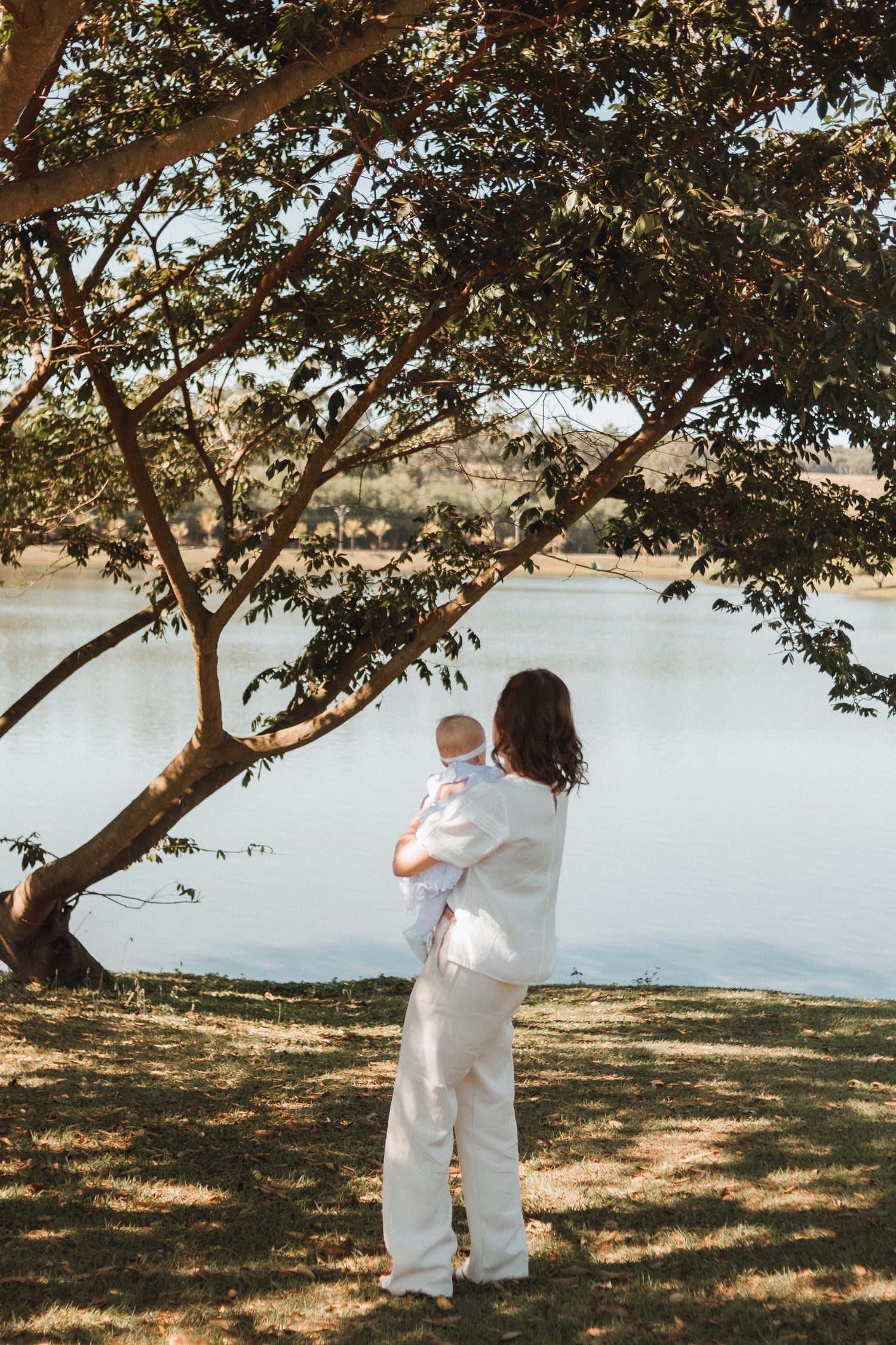 Mother in white outfit holding baby by a serene lake under a large tree on a sunny day.