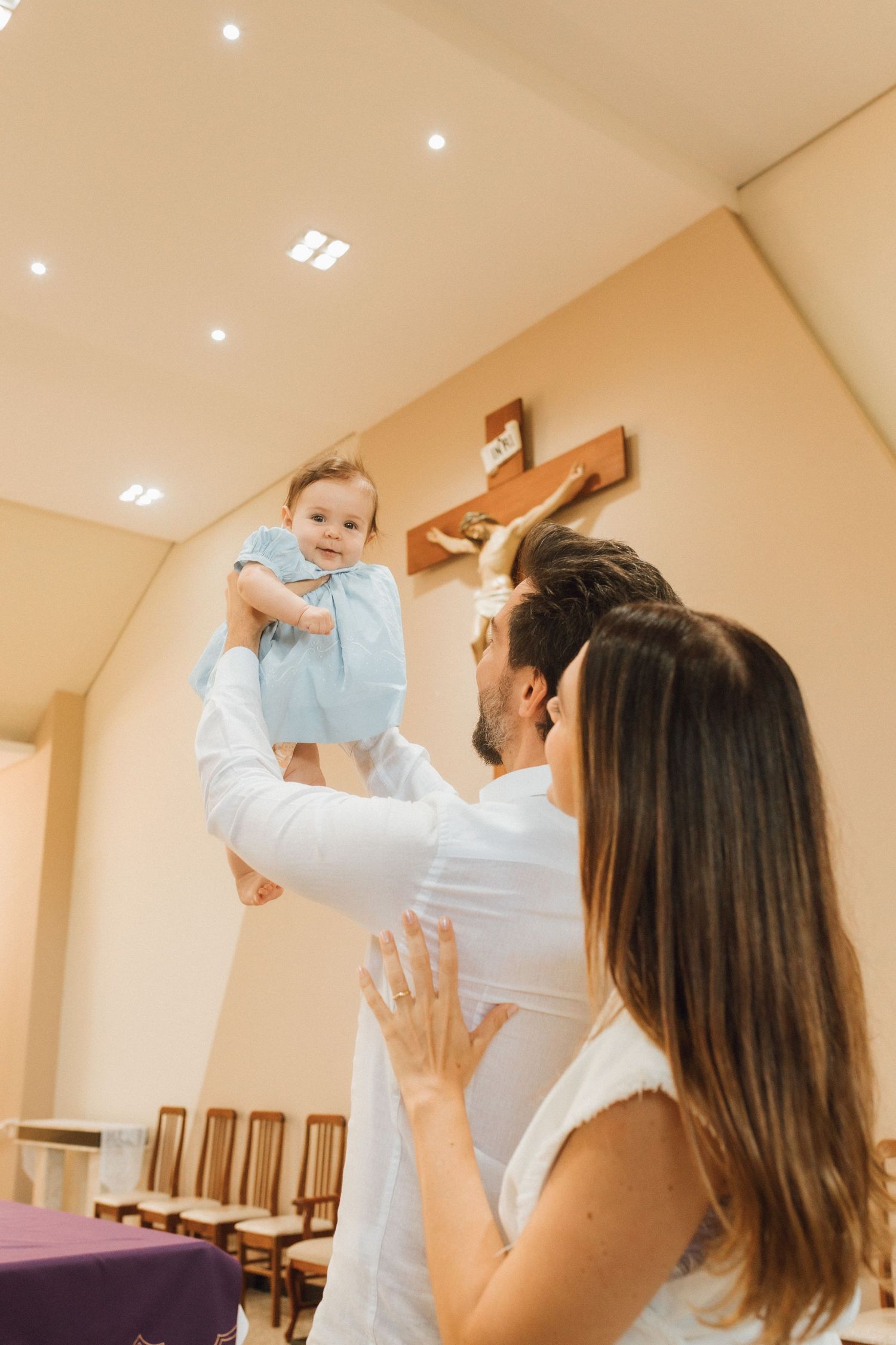 Parents joyfully lift smiling baby in blue dress inside Catholic church near crucifix.