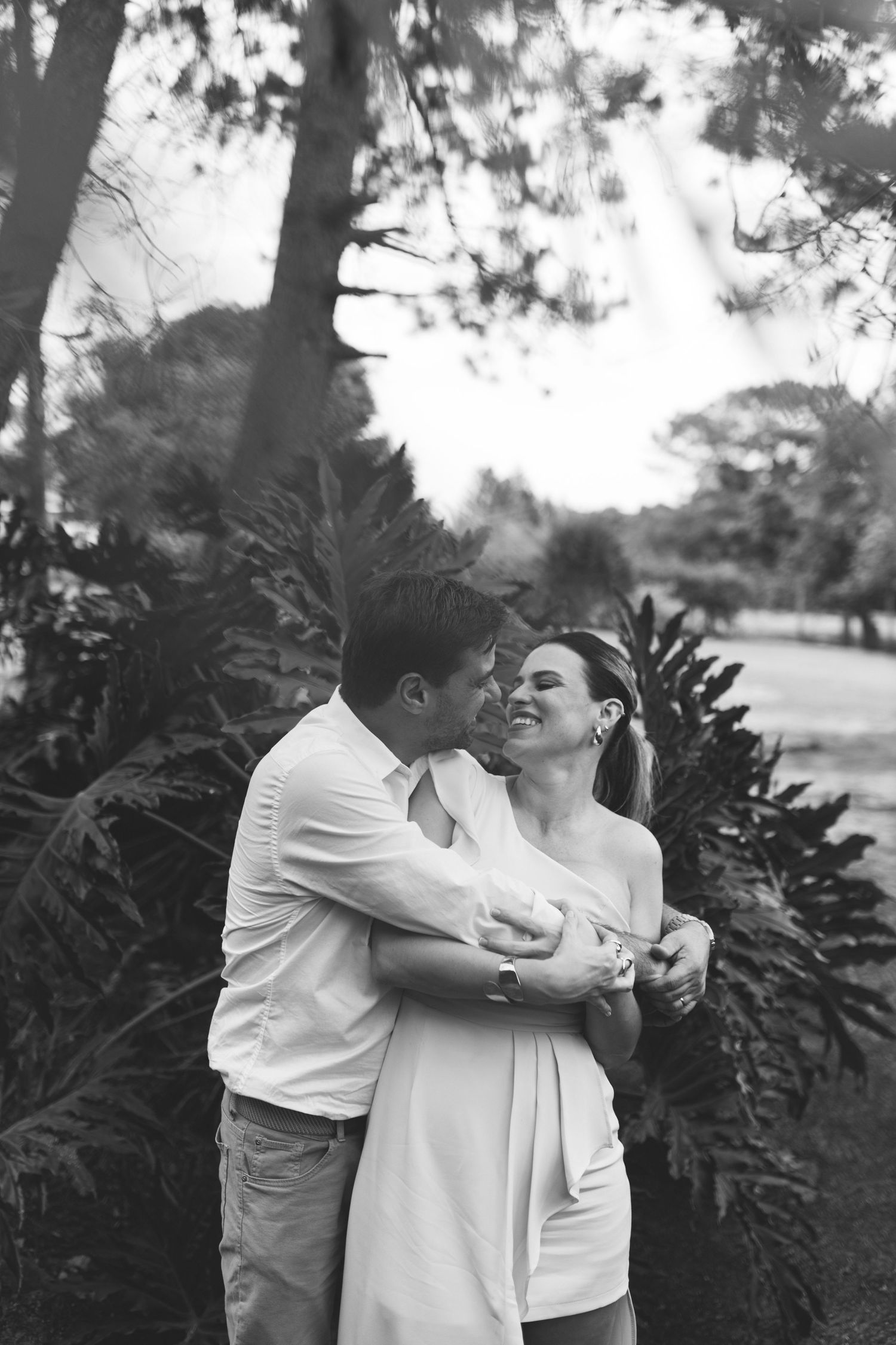 Black and white photo of a couple embracing and laughing together outdoors among lush tropical trees and ferns.
