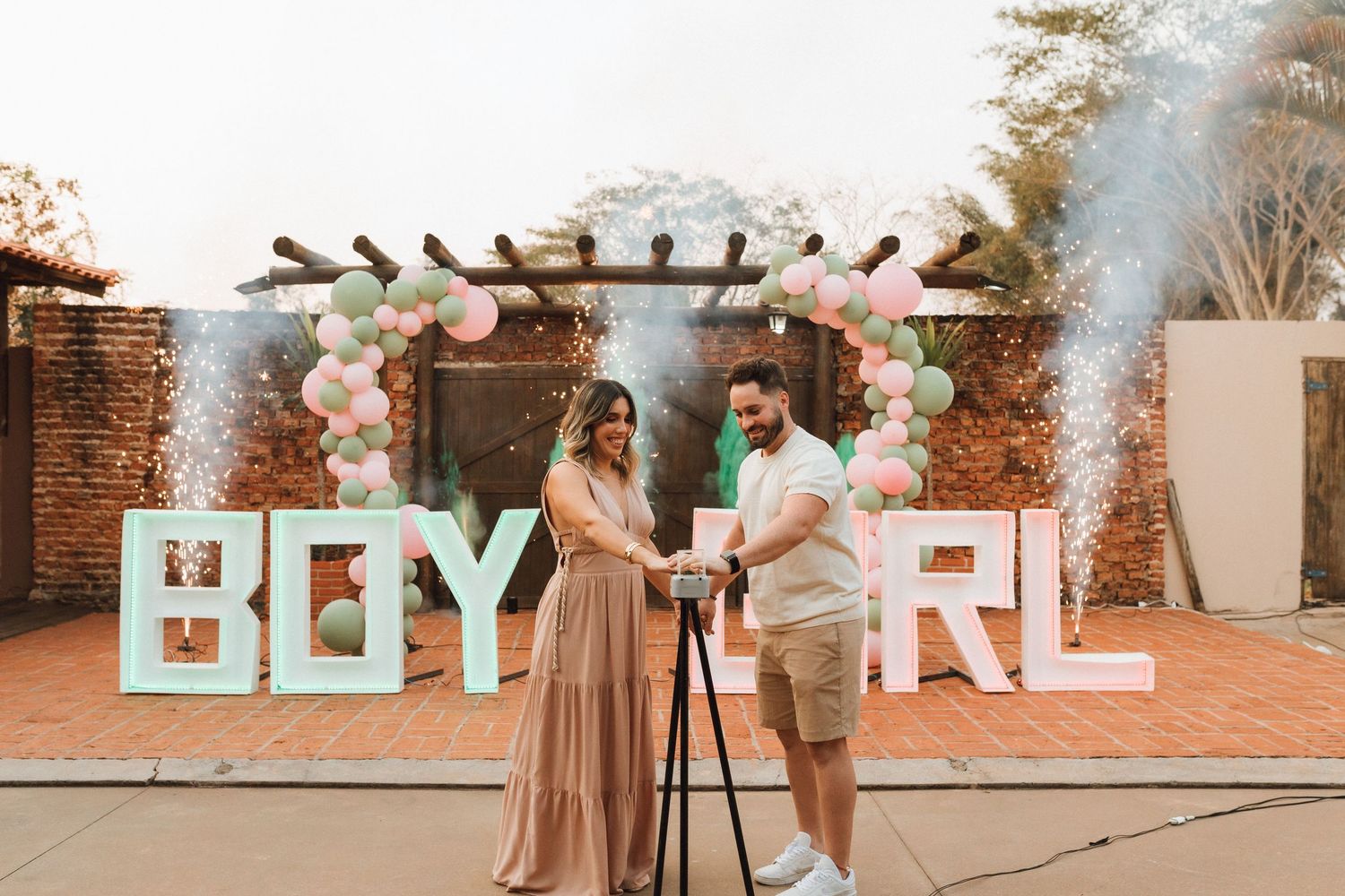 Couple cutting cake at gender reveal party with BOY and GIRL signs, balloons, and colorful smoke outdoors.