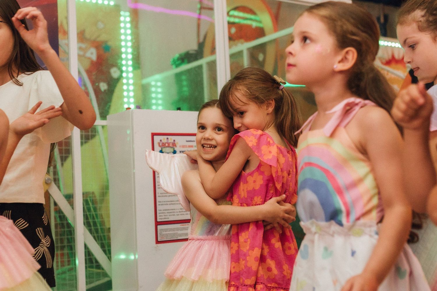 Young girls in colorful dresses hugging and laughing at a vibrant indoor children's party venue.