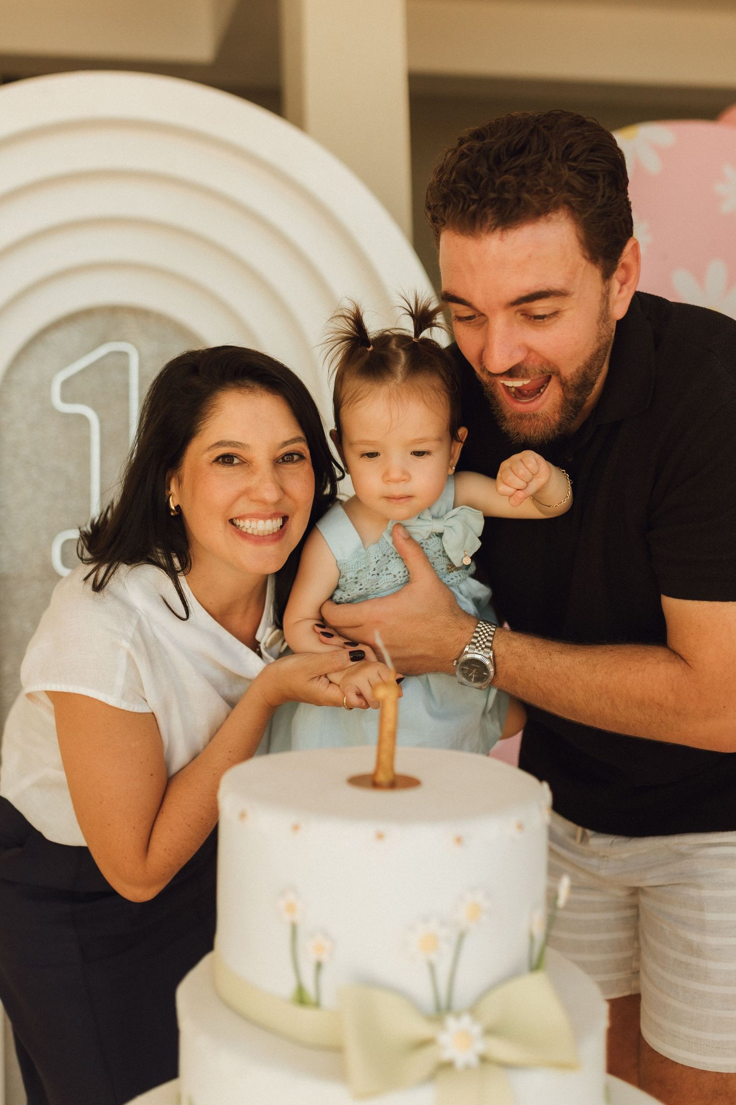 Family celebrating baby girl’s first birthday with a white floral cake and number one candle at a pastel party.