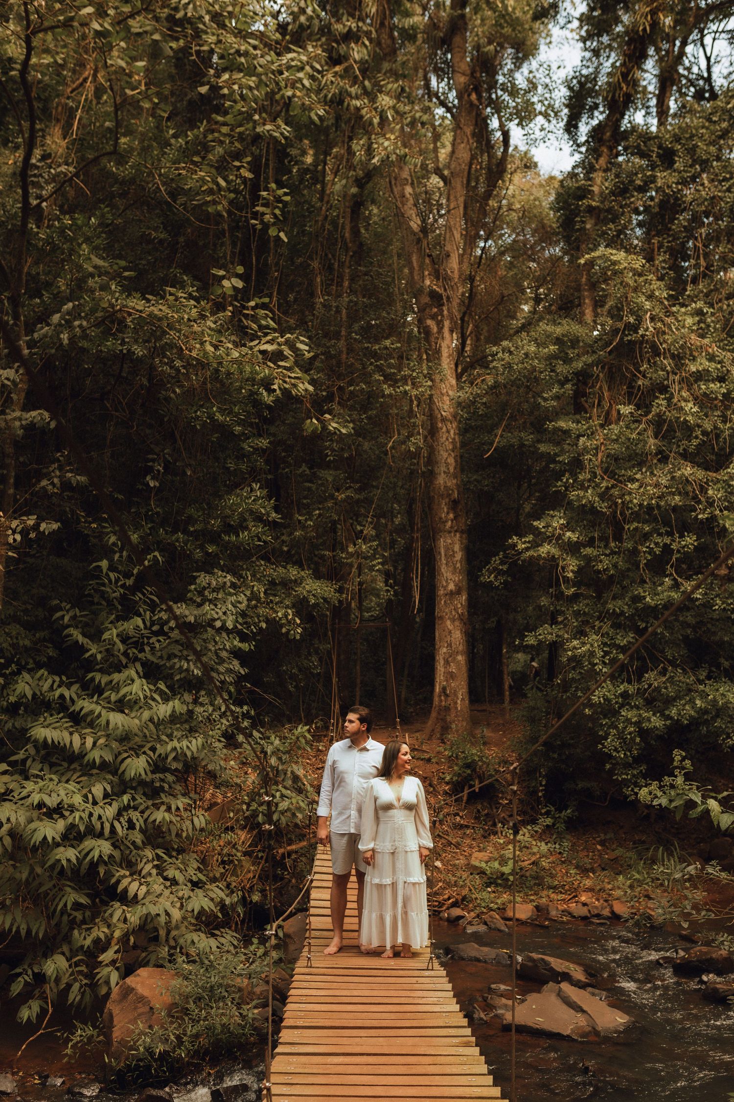 Couple in white attire standing on a wooden bridge surrounded by lush green forest trees.