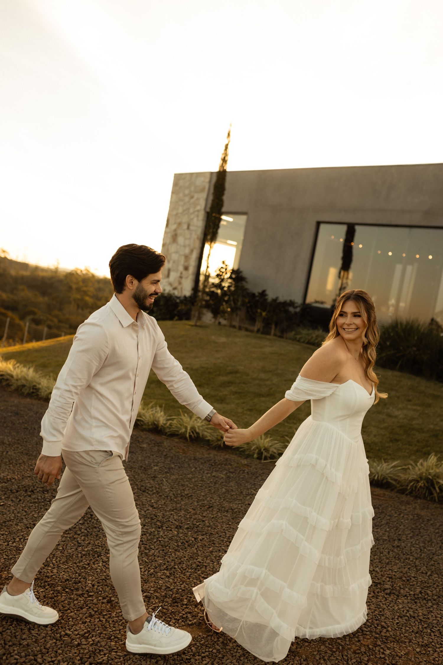 Couple in wedding attire holding hands at sunset, bride in white off-shoulder gown, groom in beige suit walking outdoors.