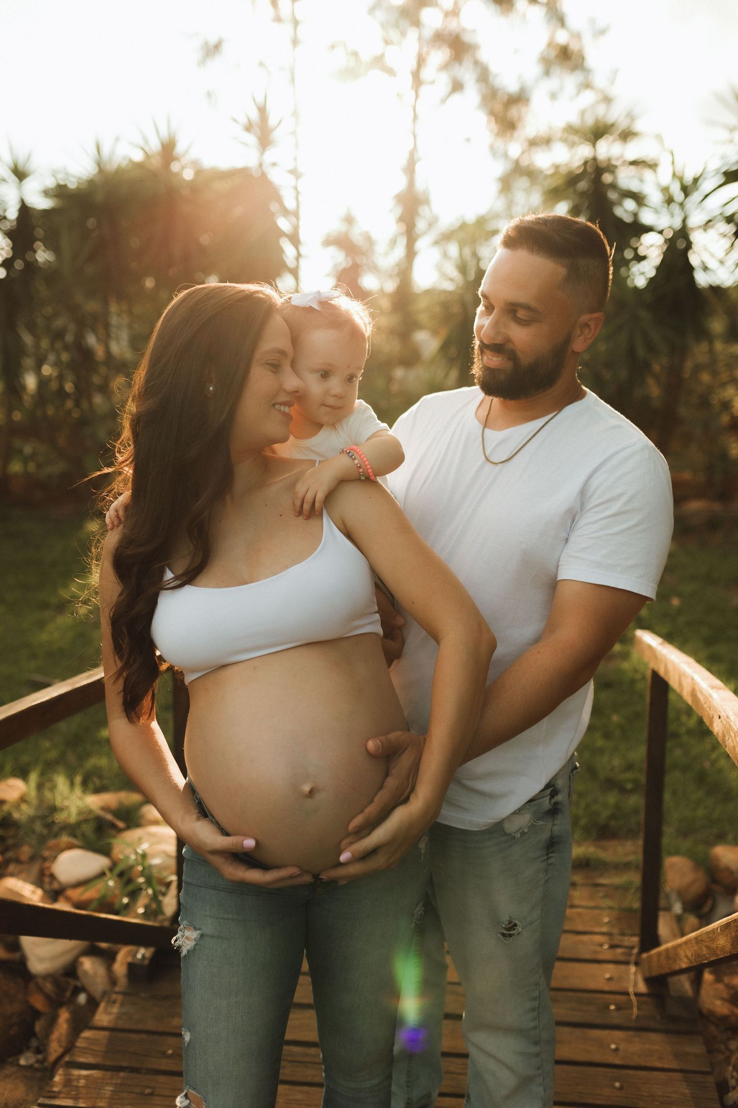 Pregnant woman and man holding toddler pose together on wooden bridge at golden hour sunset outdoors.