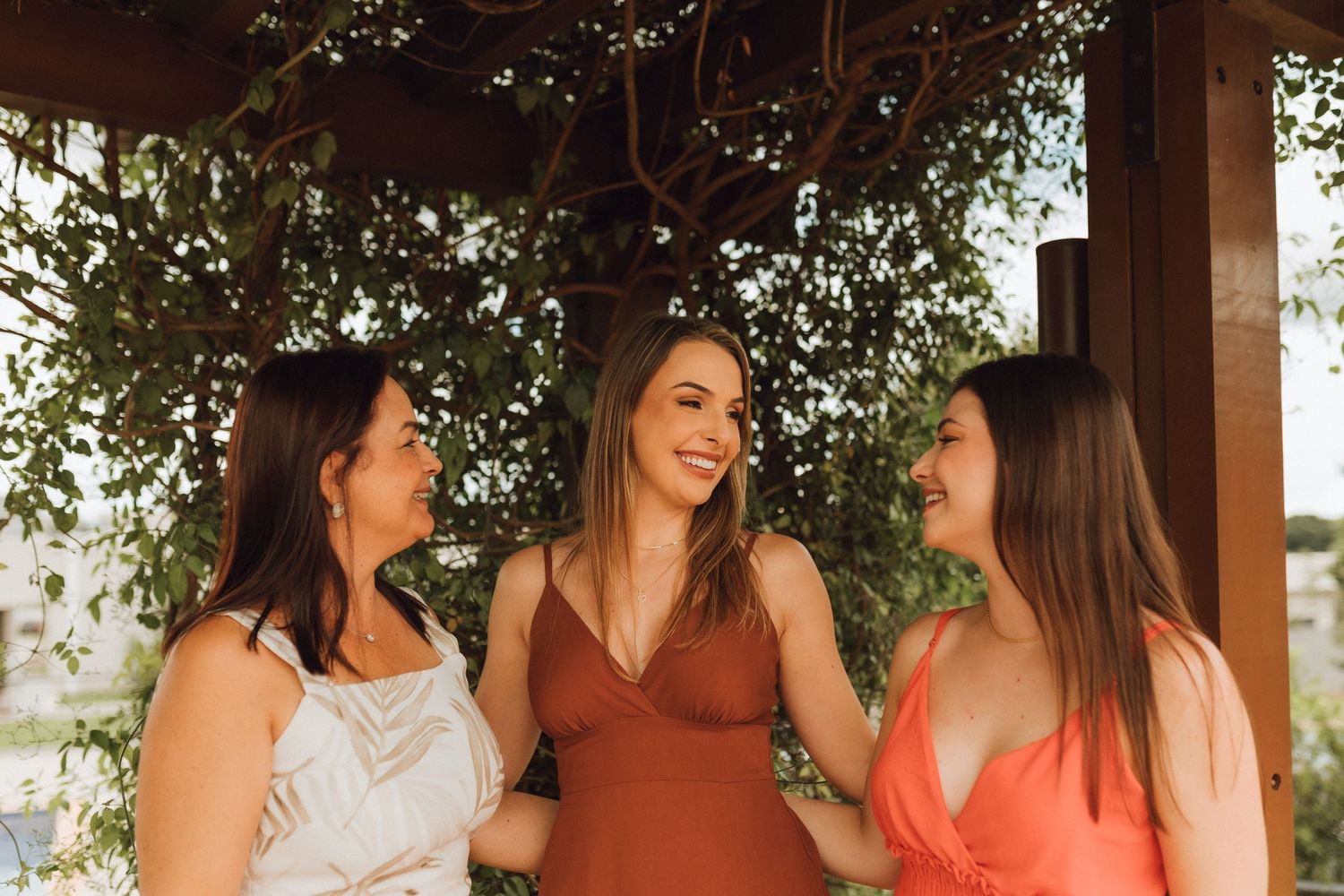 Three smiling women standing together under a wooden pergola covered in green vines outdoors.