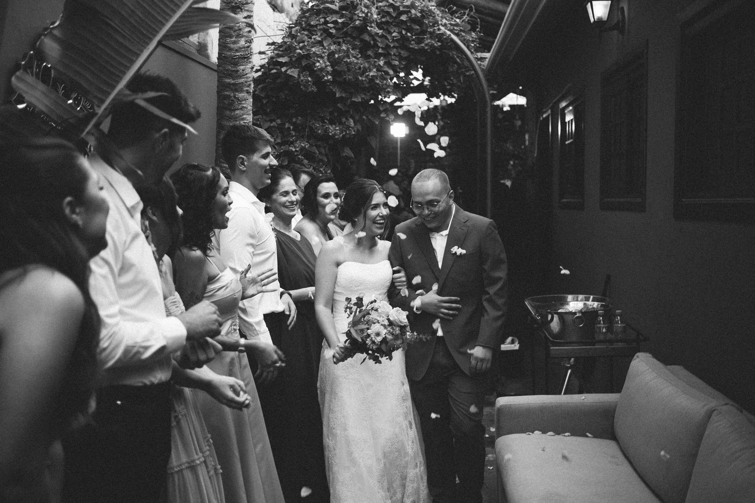 Black and white wedding photo of bride and groom walking through guests throwing confetti at outdoor reception.