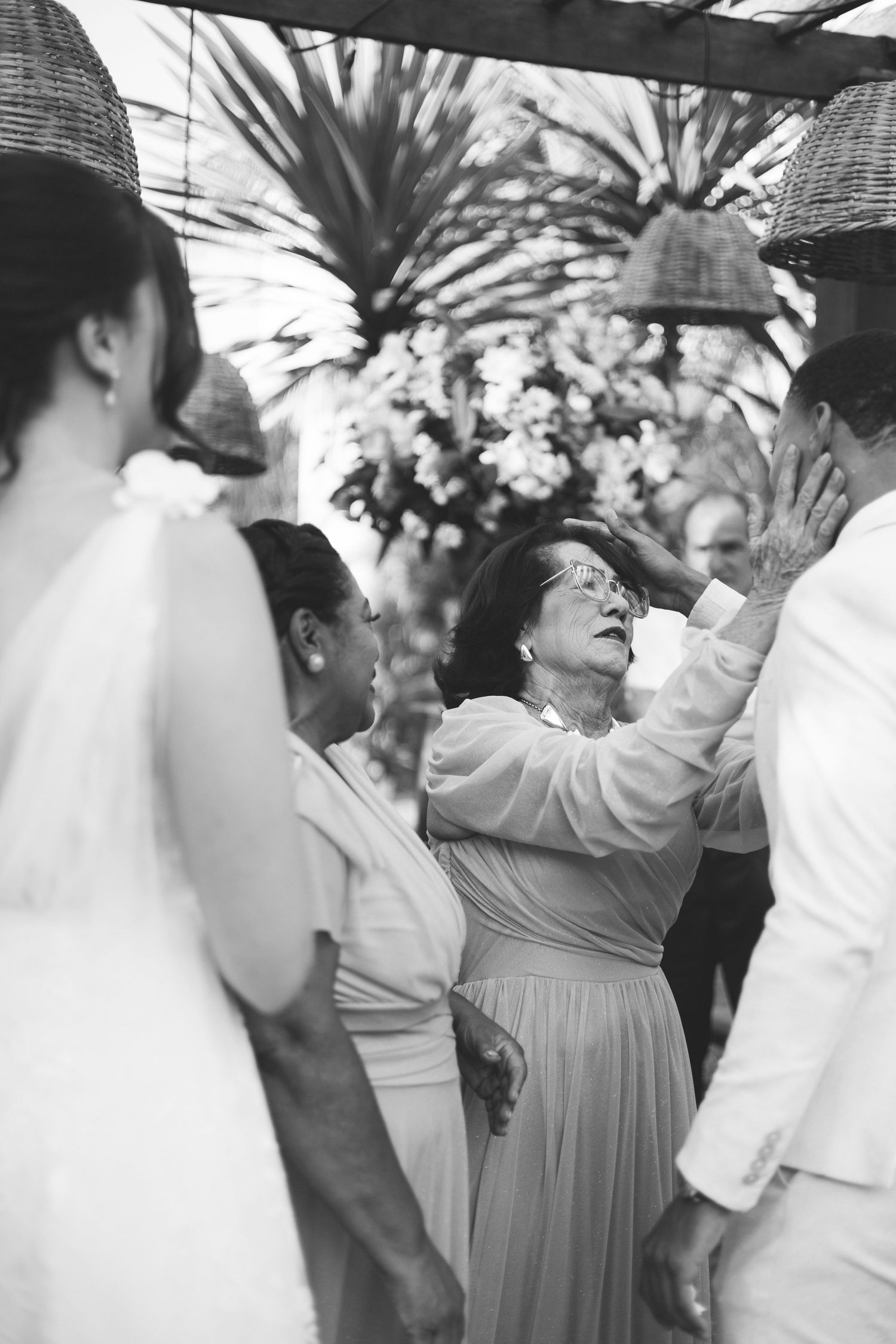 Elderly woman reaches up joyfully to touch a wedding couple during outdoor celebration.