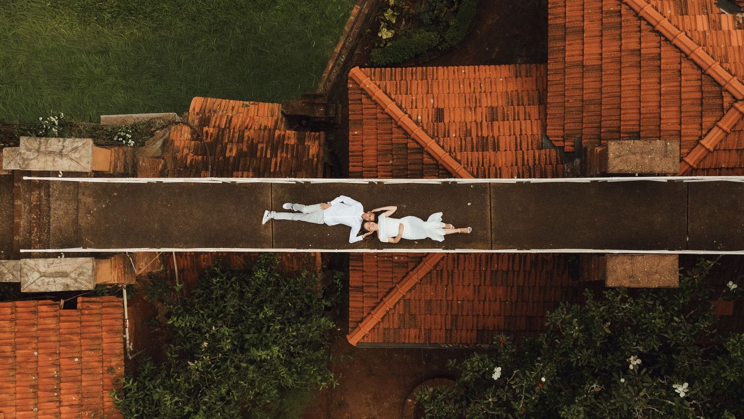 Aerial view of a couple lying on a narrow path between terracotta rooftops, dressed in white.