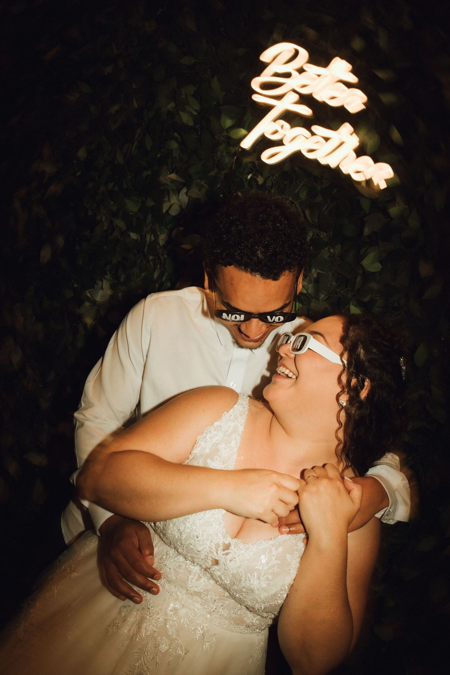 Couple embraces under a glowing Better Together neon sign surrounded by lush greenery at night.