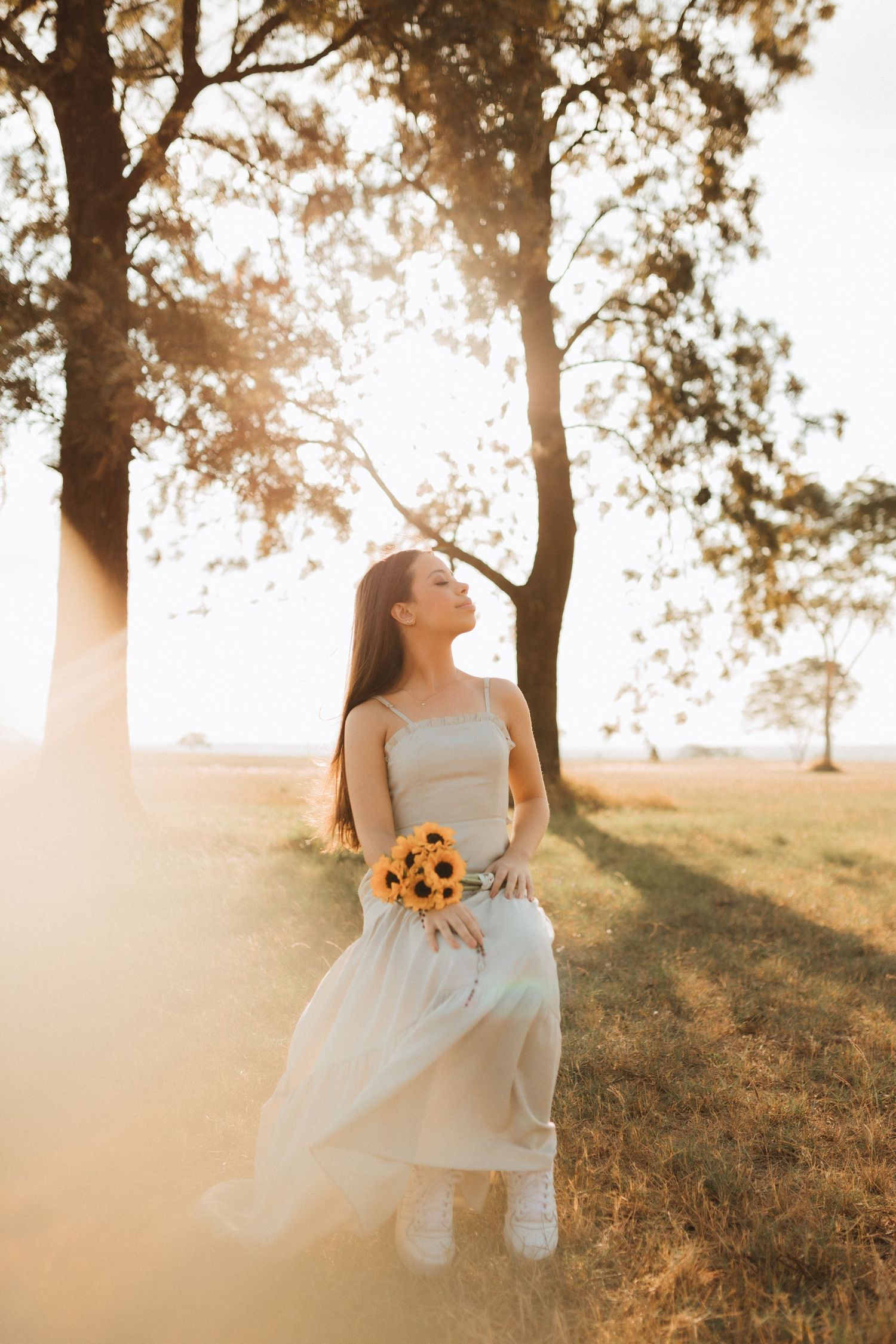 Young woman in light dress holding sunflowers, standing under trees in golden sunset light on a grassy field.