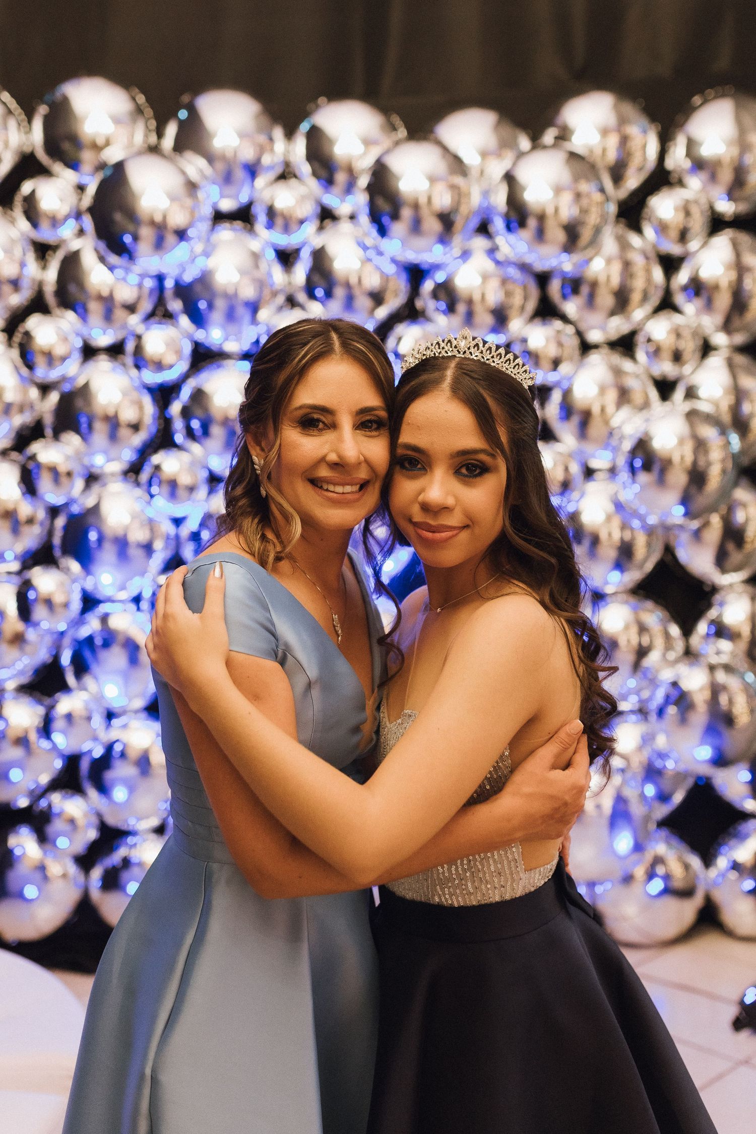 Two women embrace and smile in front of silver balloon wall, one wearing a tiara and formal gown.