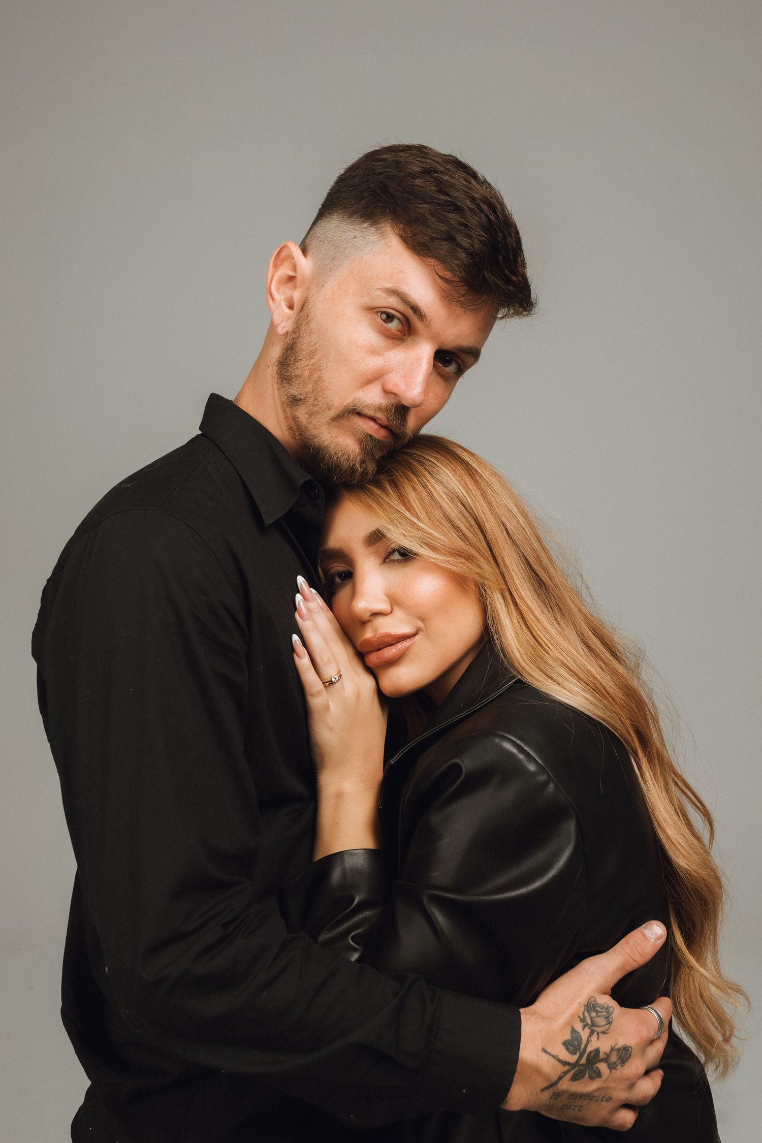 A couple in black outfits embrace tenderly, the man with tattoos holding the blonde woman close against a gray background.