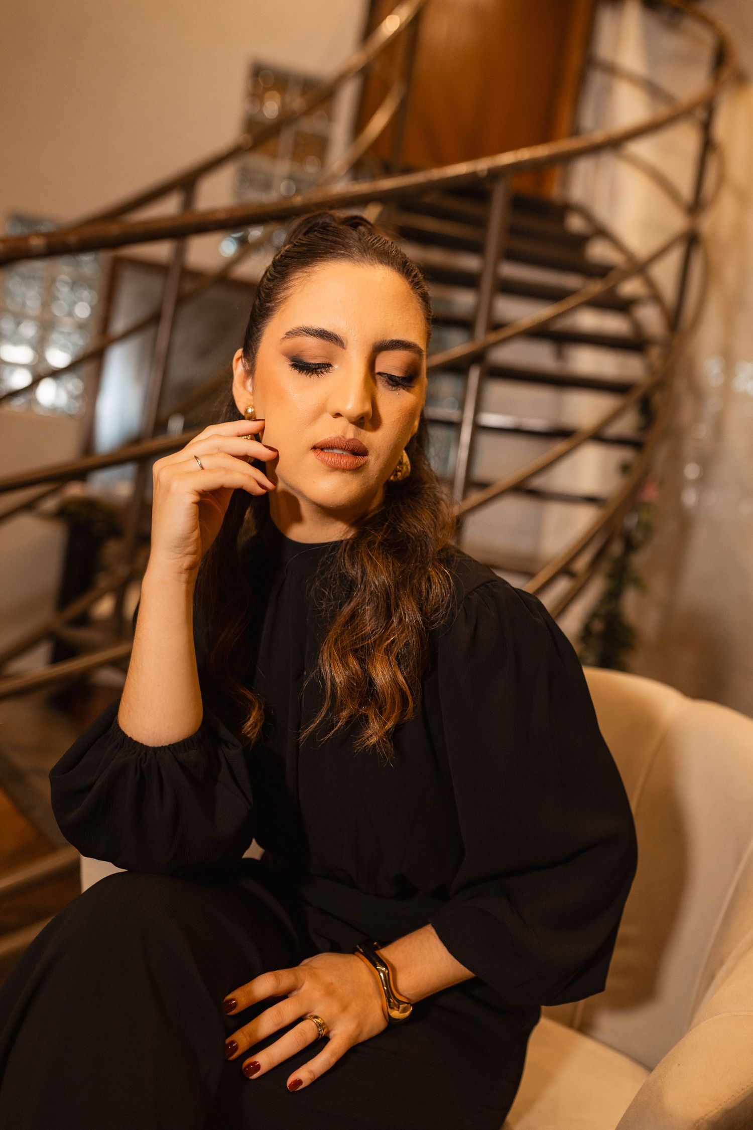 Elegant woman in black outfit sitting gracefully near spiral staircase, wearing gold jewelry with smoky eye makeup.