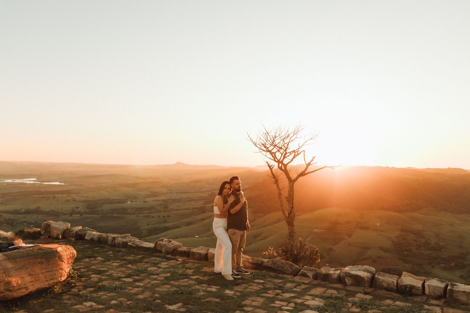 Couple embracing at golden sunset on scenic hilltop overlook with lush green valley and bare tree backdrop.
