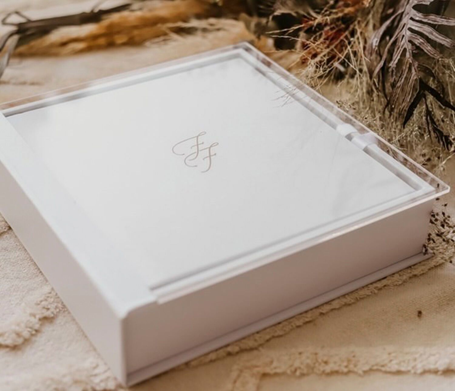 Elegant white keepsake box with gold monogram initials, displayed on a neutral textured surface with dried botanicals.