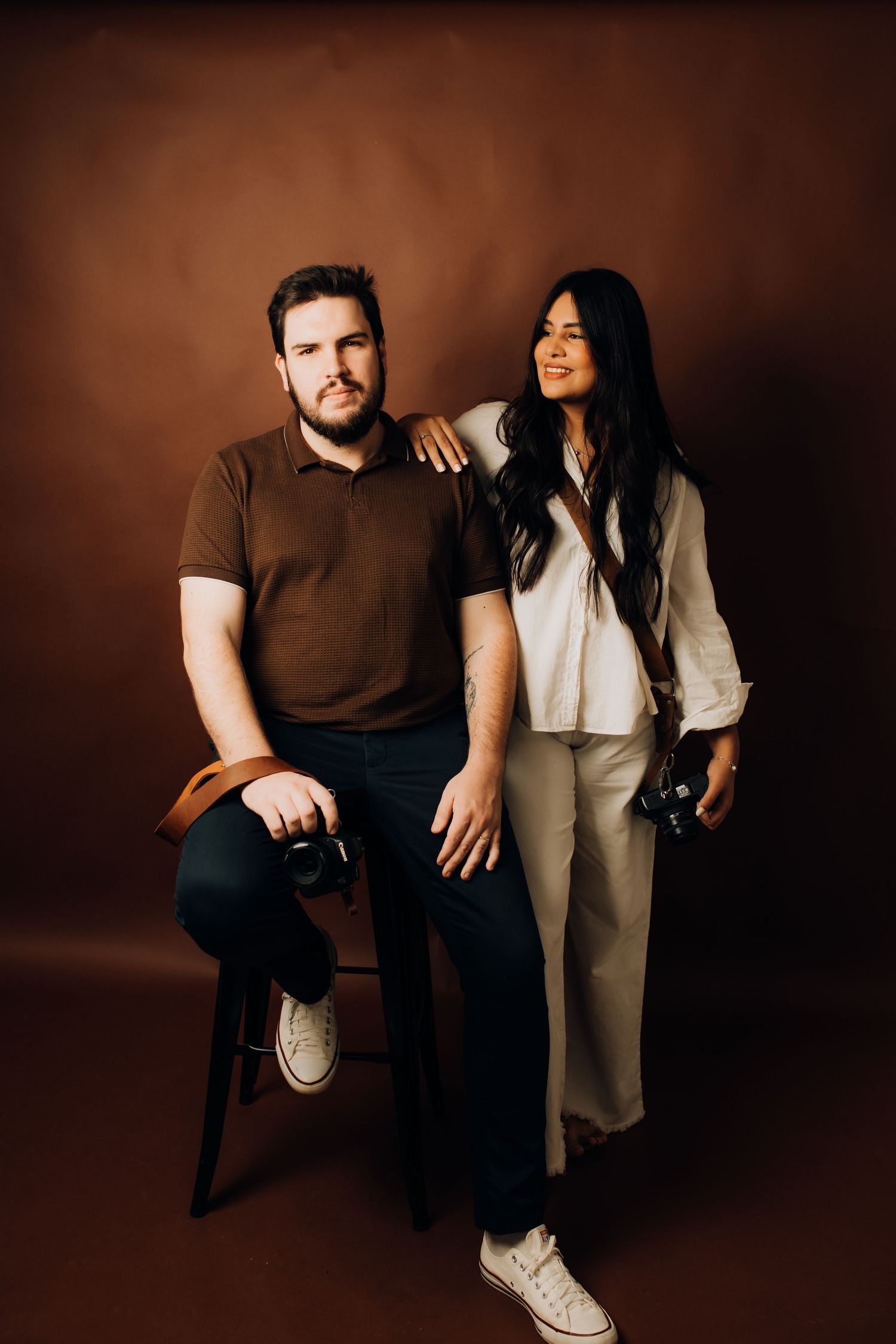 A man and woman pose in a studio, both holding cameras, against a warm brown background.