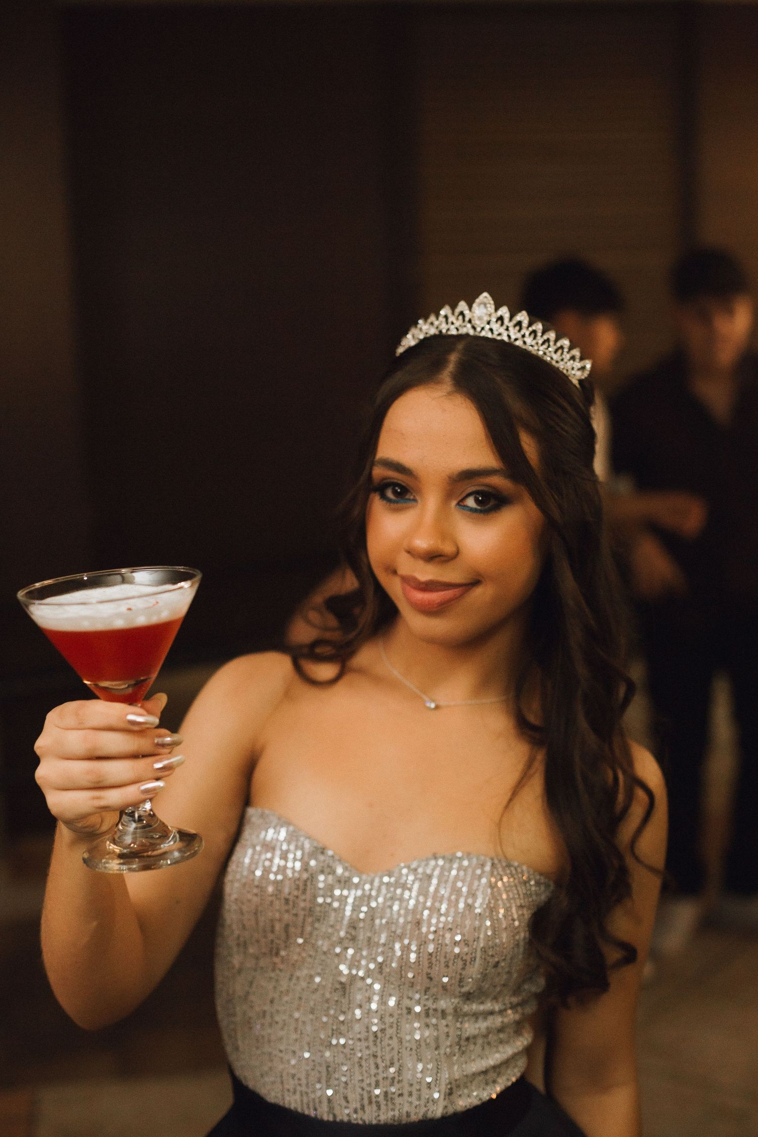 Young woman in silver sequin strapless gown and tiara holding red cocktail at quinceañera celebration.
