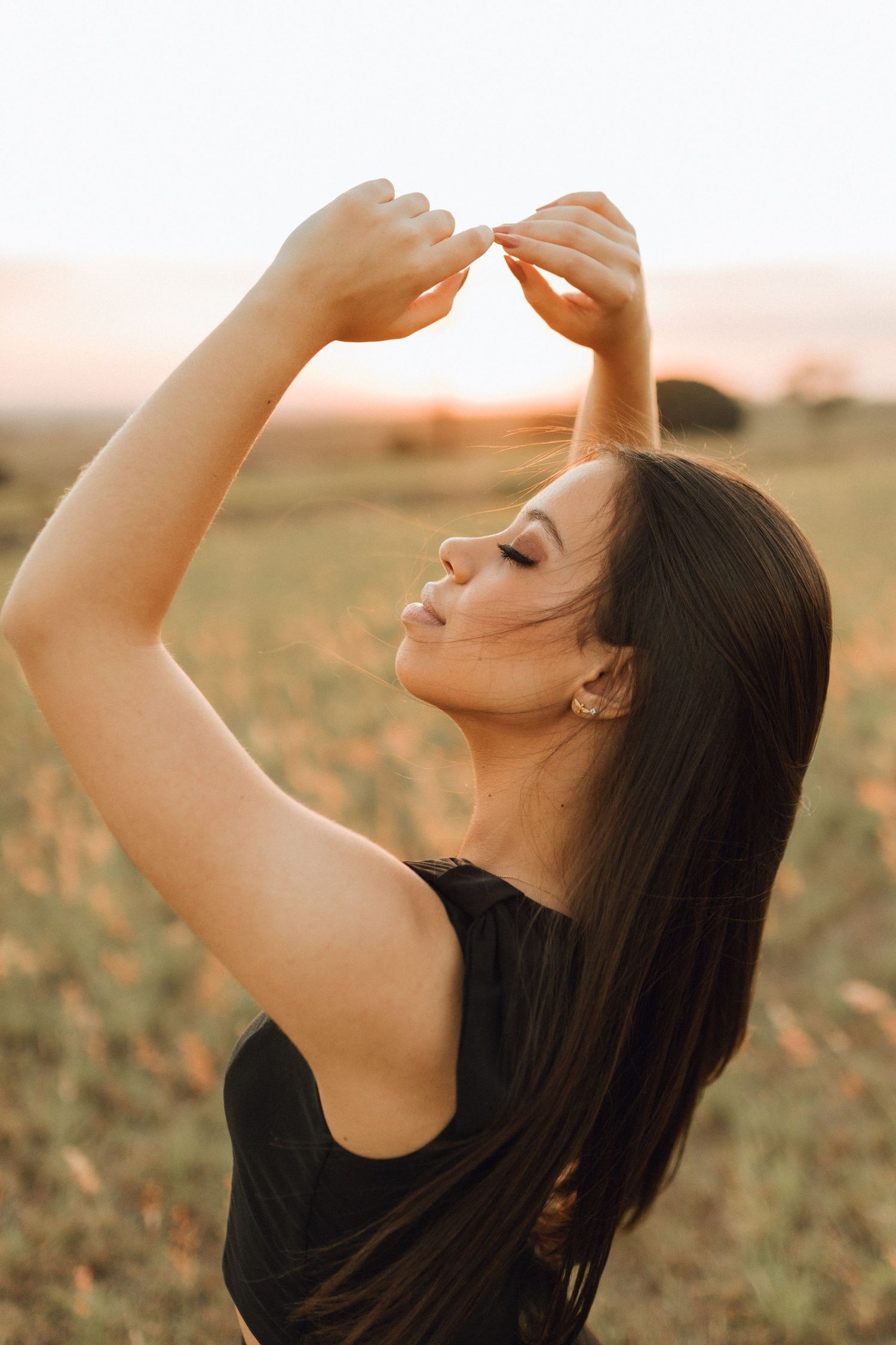 Young woman with long dark hair poses at sunset in a field, arms raised, eyes closed, wearing a black top.