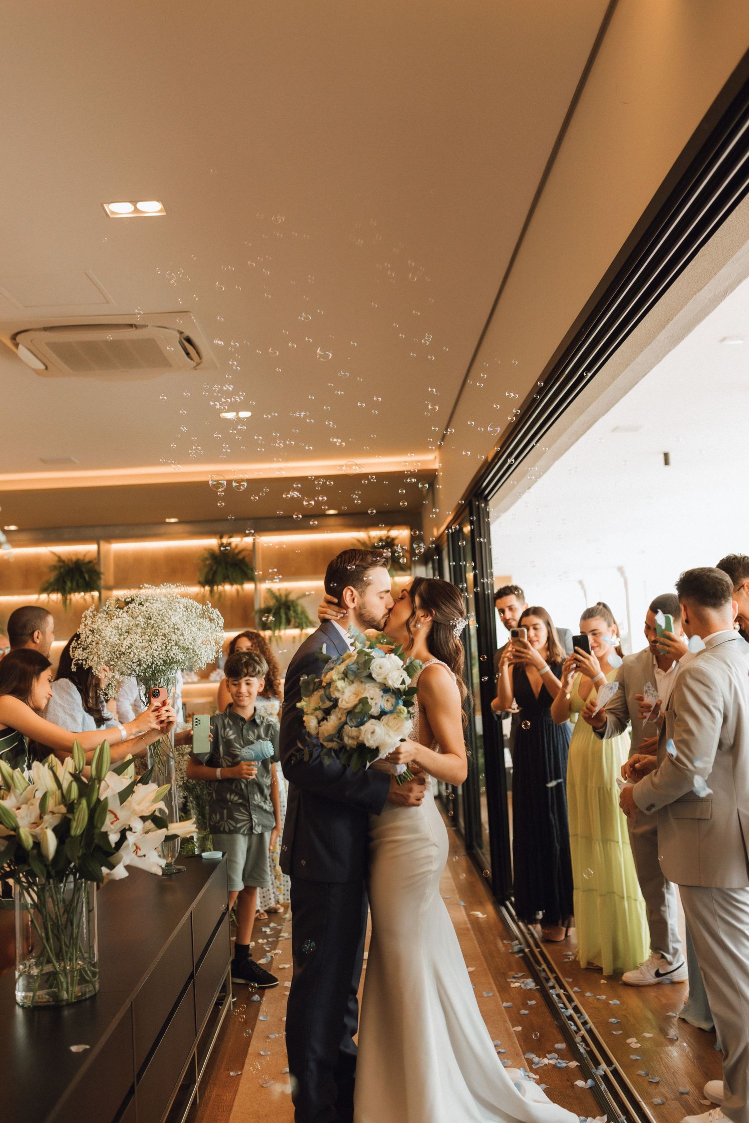 Newlyweds share a first kiss as guests celebrate with bubbles in a warmly lit reception venue.