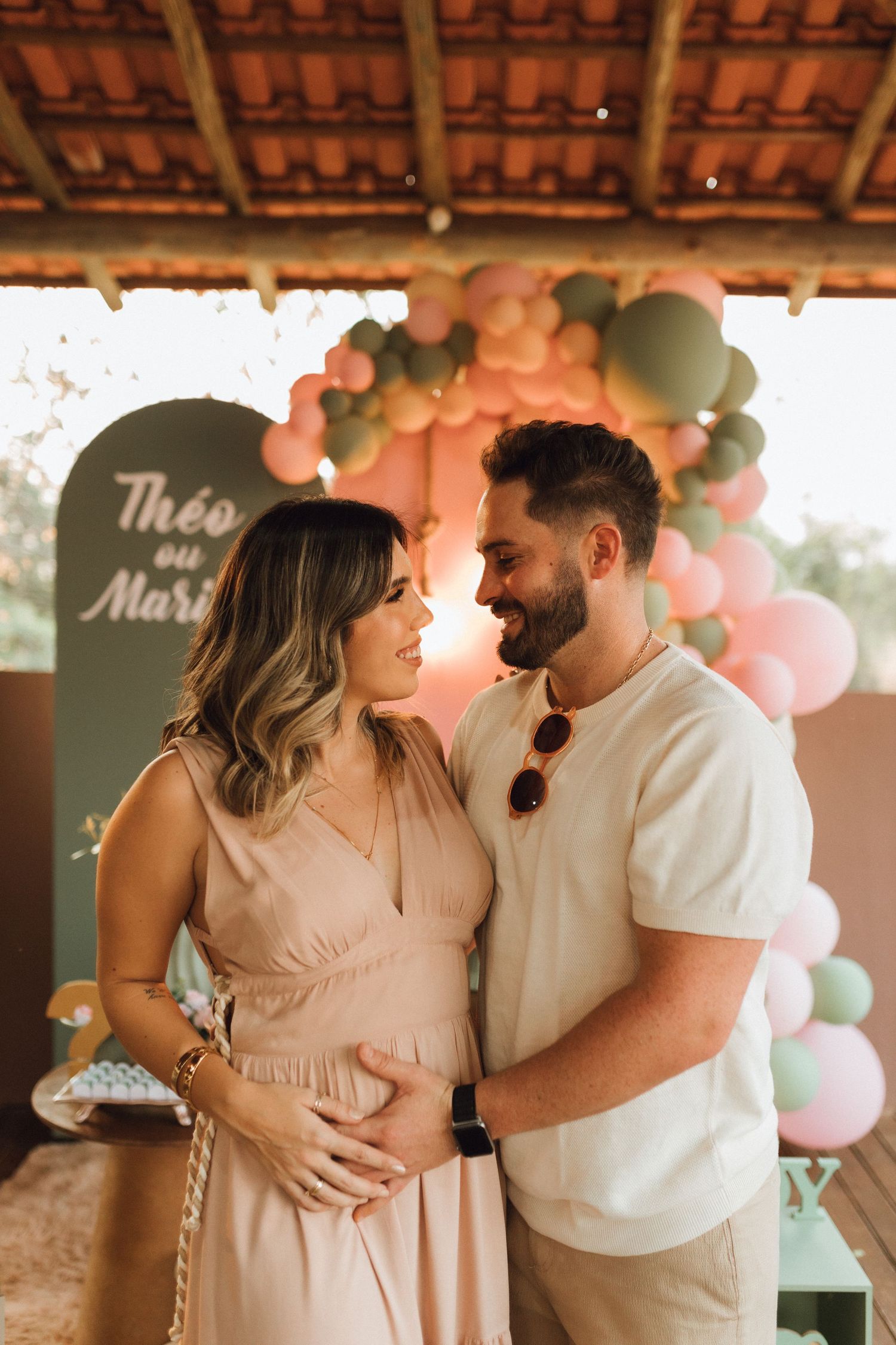 Pregnant couple tenderly embracing at a gender reveal party with pink and green balloon arch backdrop.