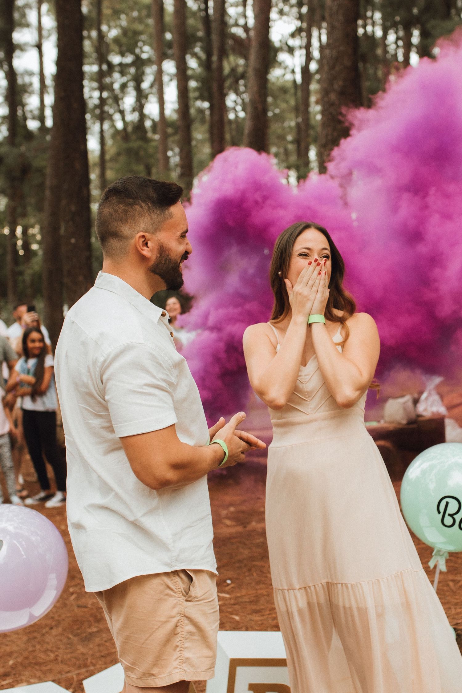 Couple reacts with joy at a pink smoke gender reveal party in a forest setting with colorful balloons.