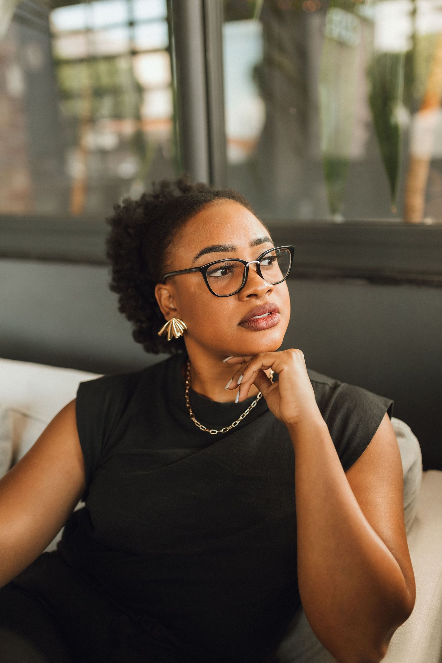 Thoughtful Black woman in glasses and black outfit sitting on couch with hand on chin.
