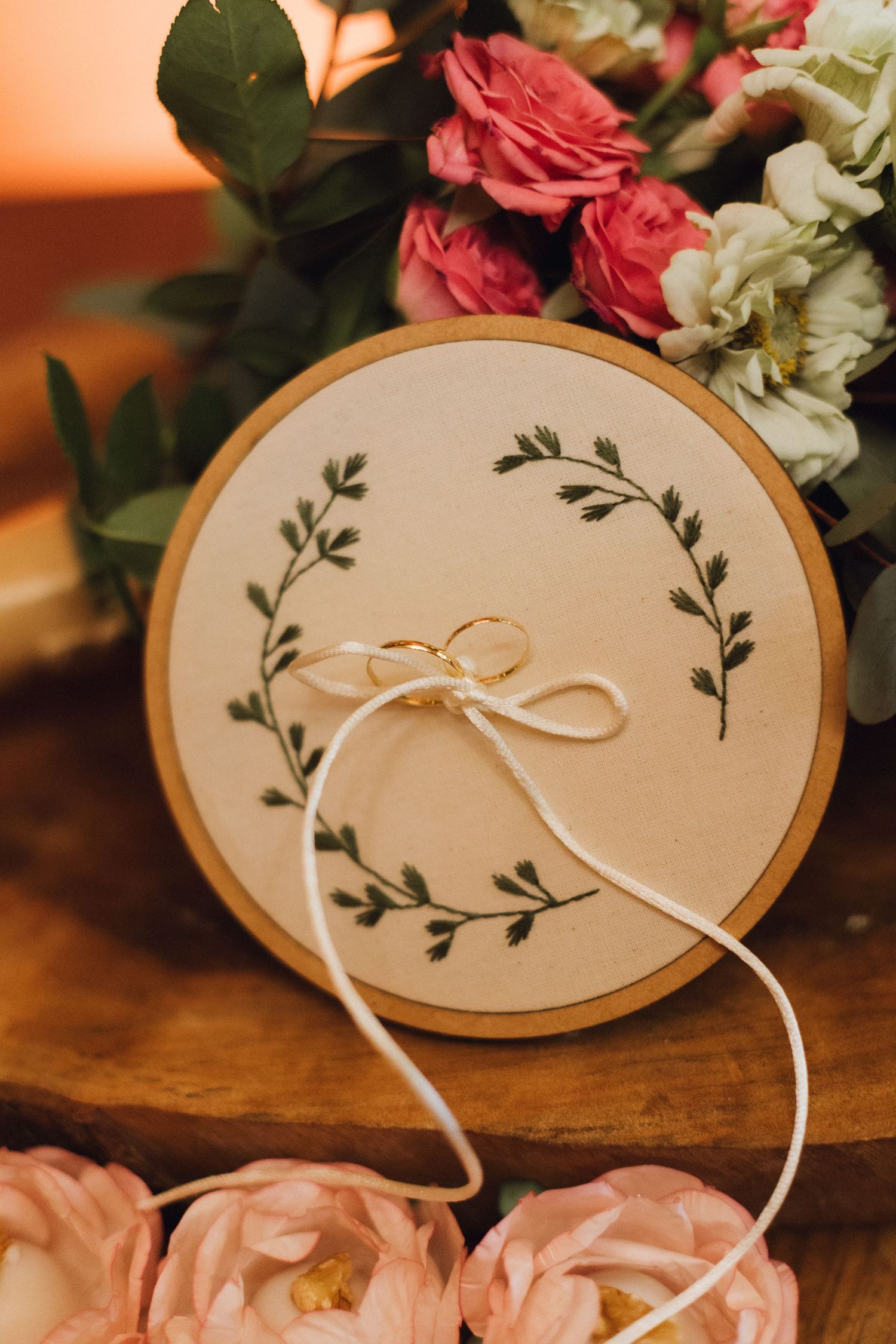 Gold wedding rings tied with ribbon on an embroidered hoop surrounded by pink flowers.