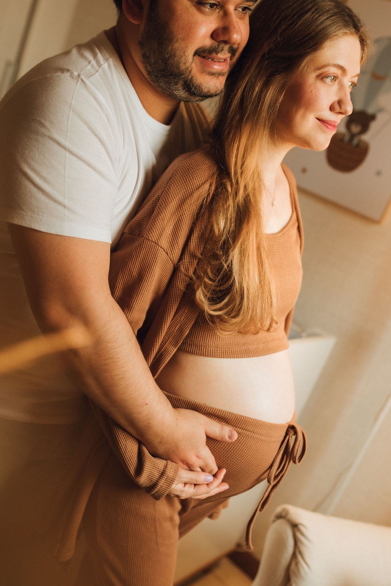 Man tenderly holds pregnant partners belly from behind in warm cozy interior setting.