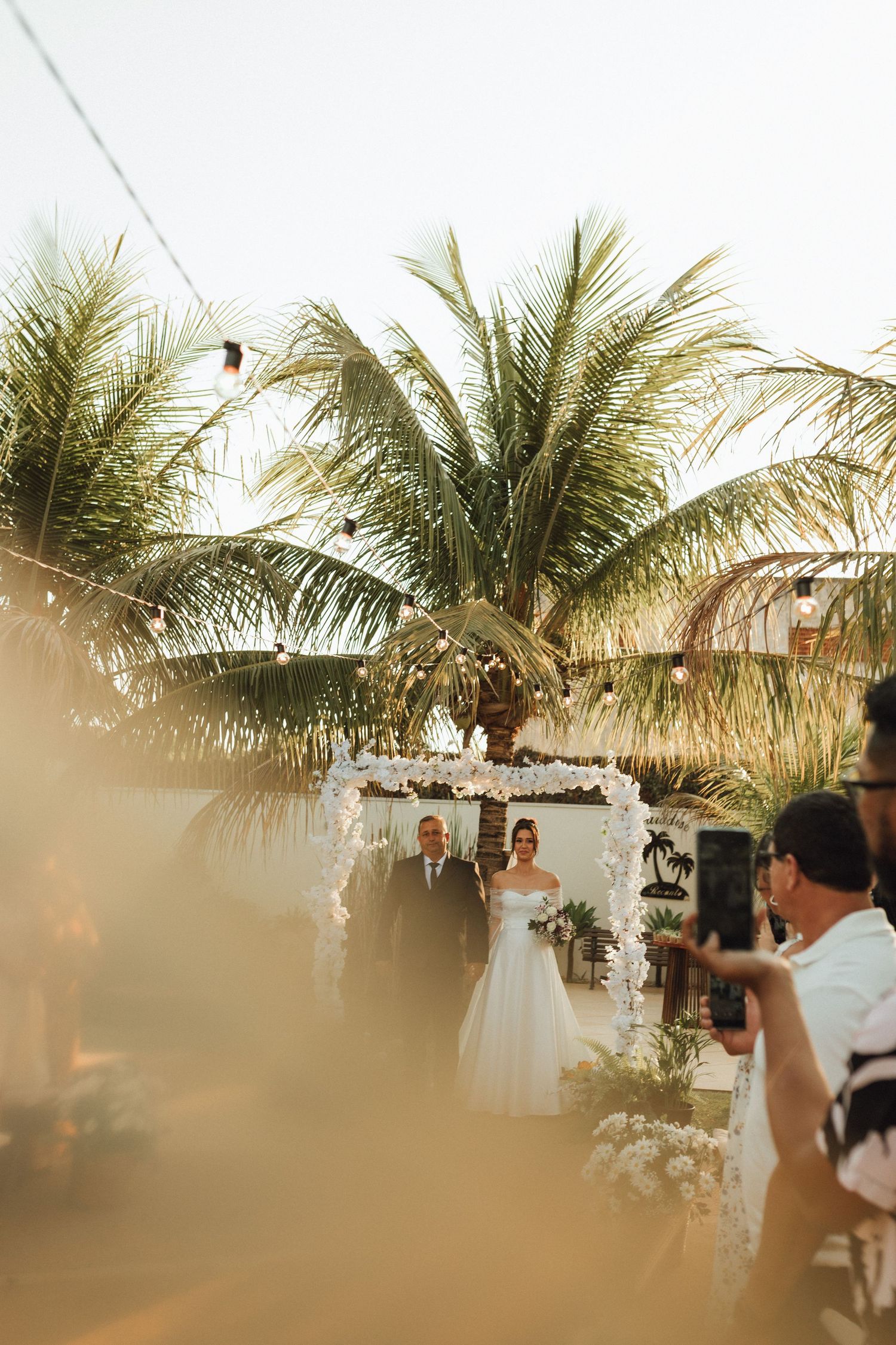 Bride walking down the aisle with escort at outdoor tropical wedding ceremony at sunset.