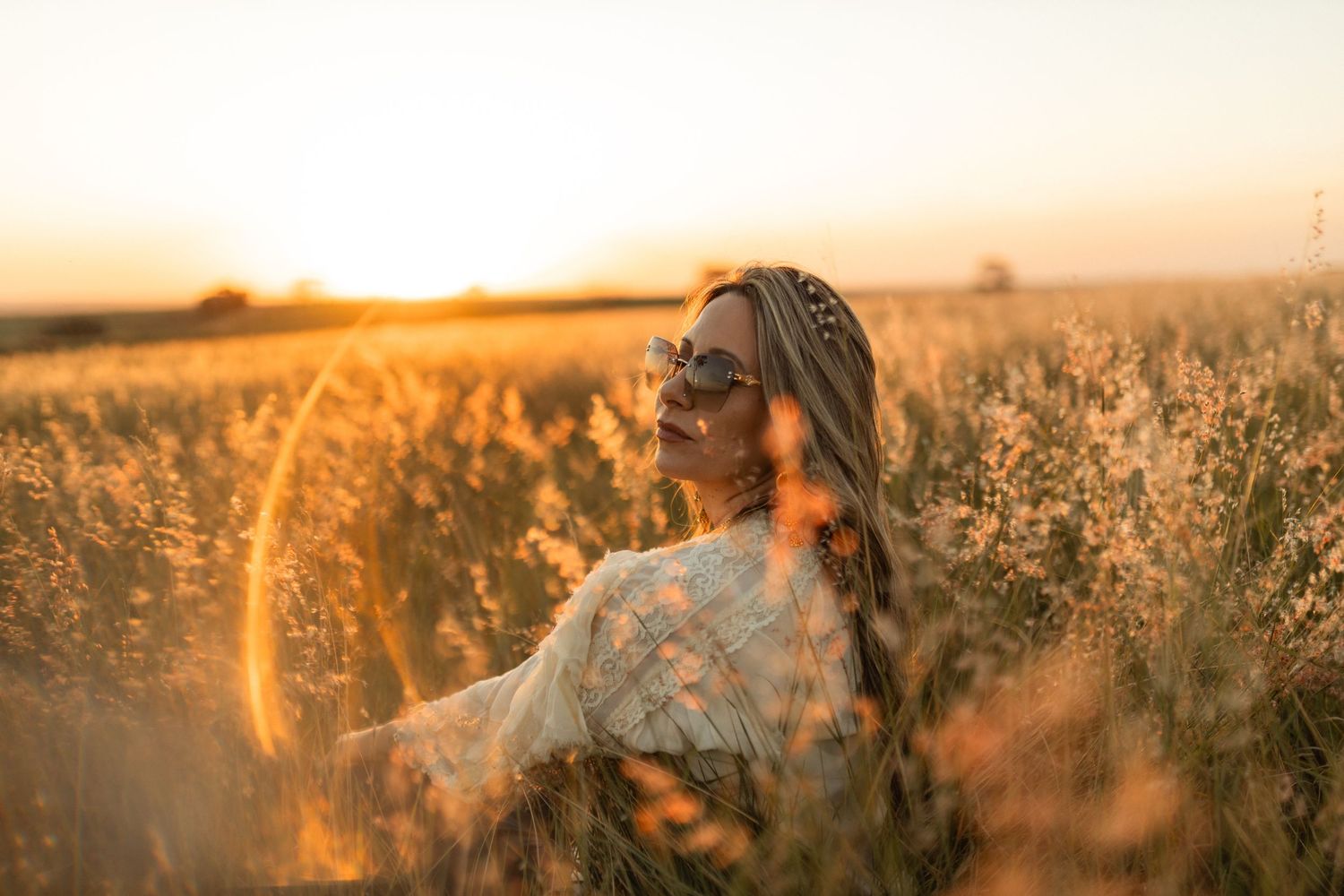 Woman in sunglasses and lace top sitting in golden wheat field at sunset with warm glowing light.