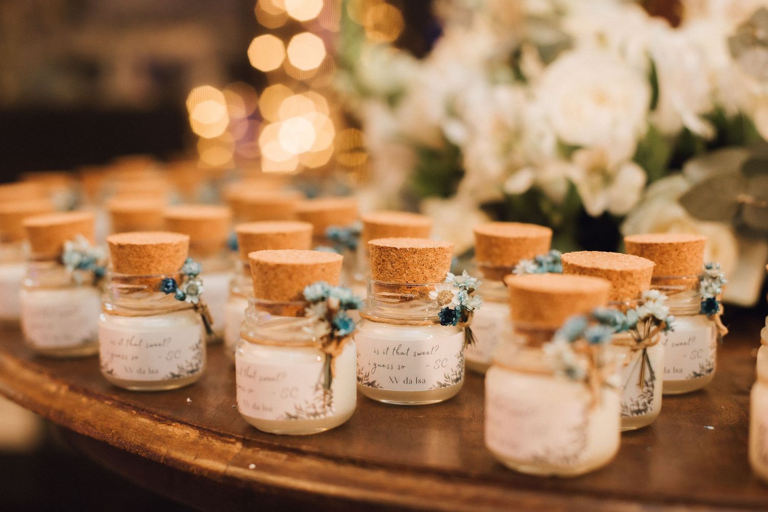 Small cork-topped glass jars with blue flowers and labels arranged as wedding favors on a wooden tray.