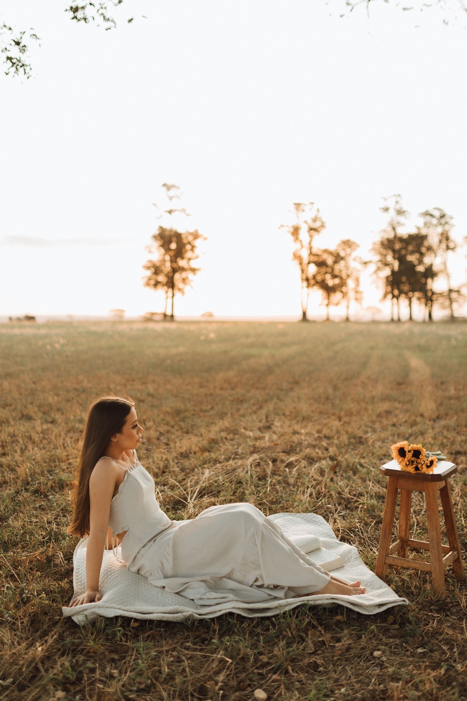 Bride in white gown sitting in golden field at sunset with sunflower bouquet on wooden stool.